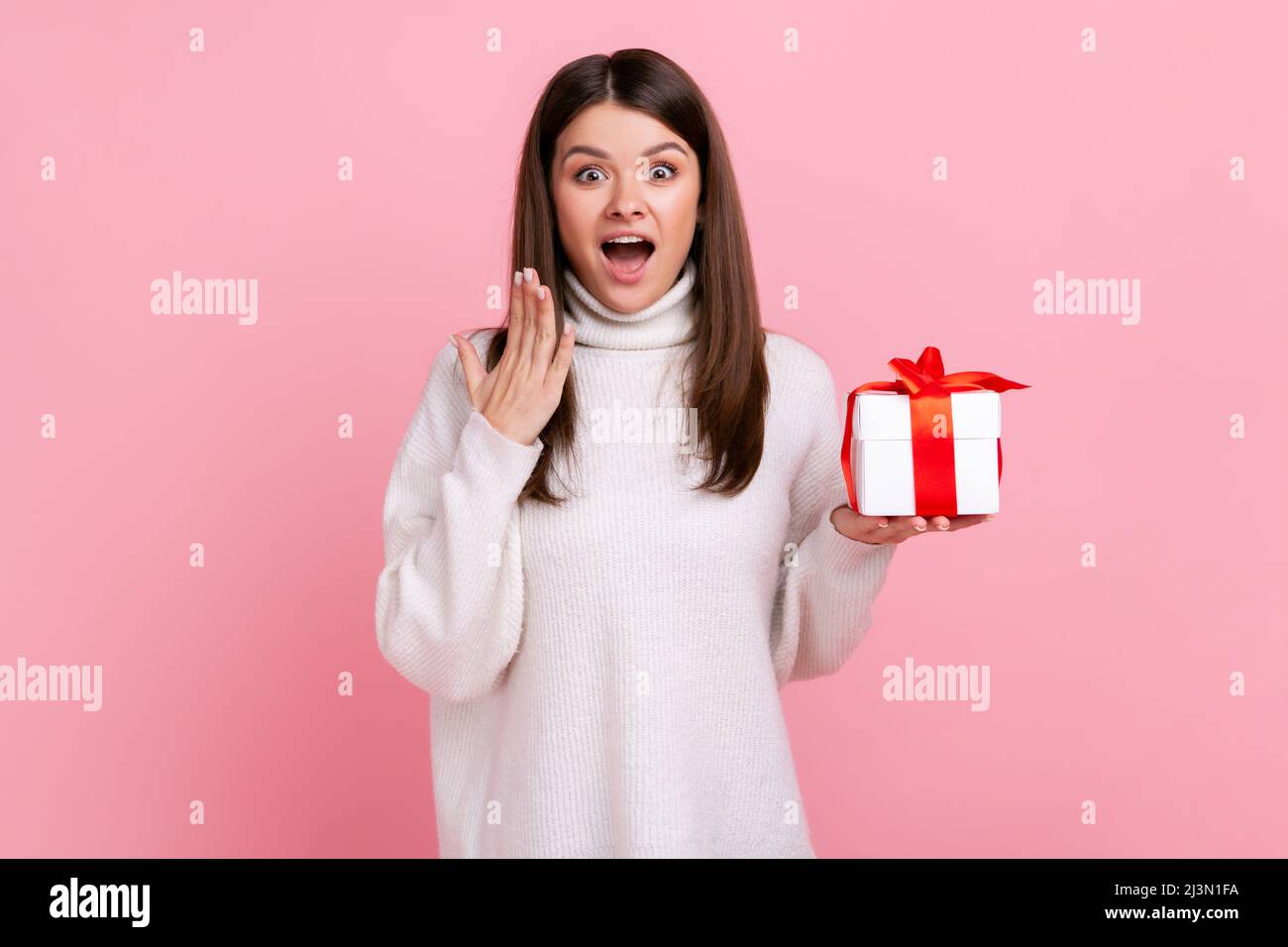 Überraschte Frau mit dunklem Haar, die eingewickelte Geschenk-Box hält, mit Erstaunen die Kamera anschaut und einen weißen Pullover im lässigen Stil trägt. Innenaufnahme des Studios isoliert auf rosa Hintergrund. Stockfoto
