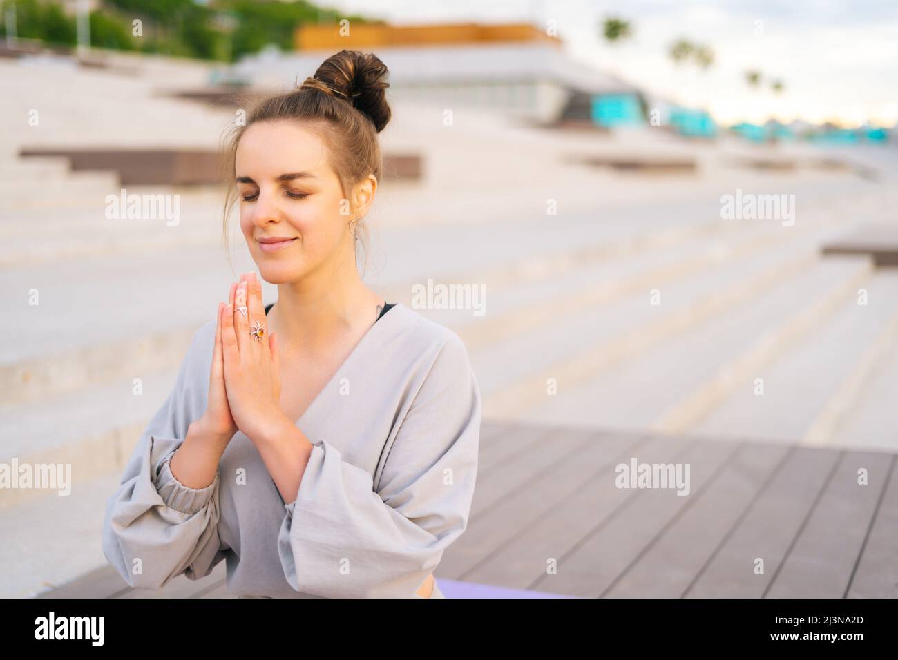 Porträt einer lächelnden kaukasischen jungen Frau, die im Stadtpark Yoga praktiziert und Namaste-Pose mit geschlossenen Augen ausführt. Stockfoto