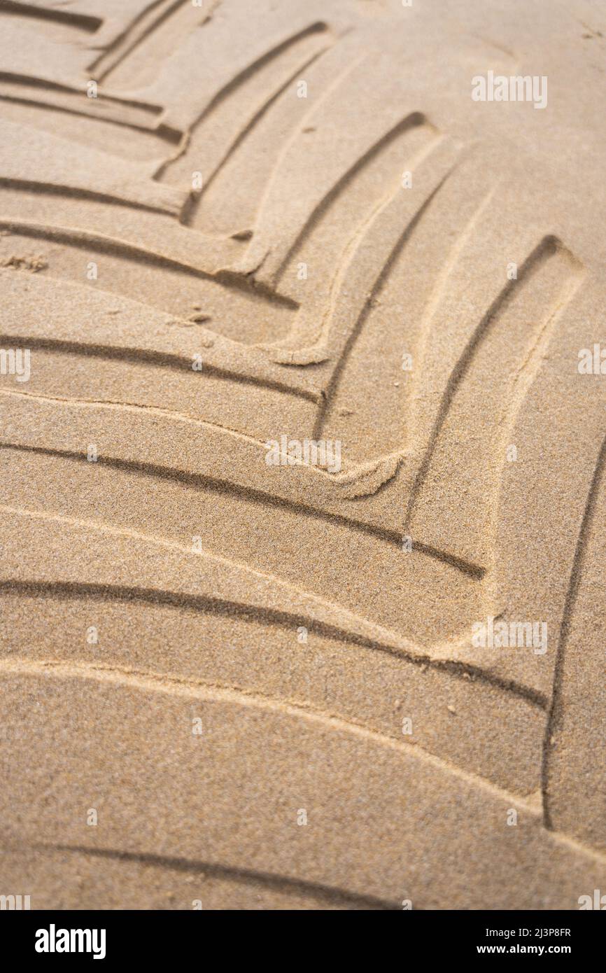 Traktorreifen-Drucke, die in den Sandstrand eingebettet sind, mit geometrischem Muster. Autoreifenspuren schließen sich am Meer mit sandiger Textur an. Stockfoto