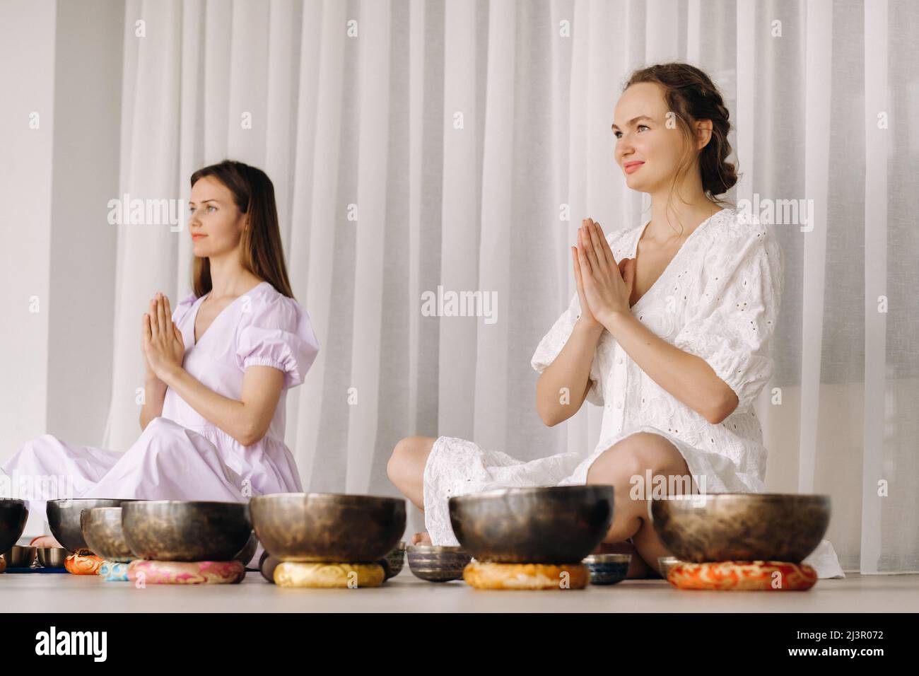 Zwei Frauen sitzen mit tibetischen Schalen in der Lotusposition vor einem Yoga-Kurs im Fitnessstudio. Stockfoto