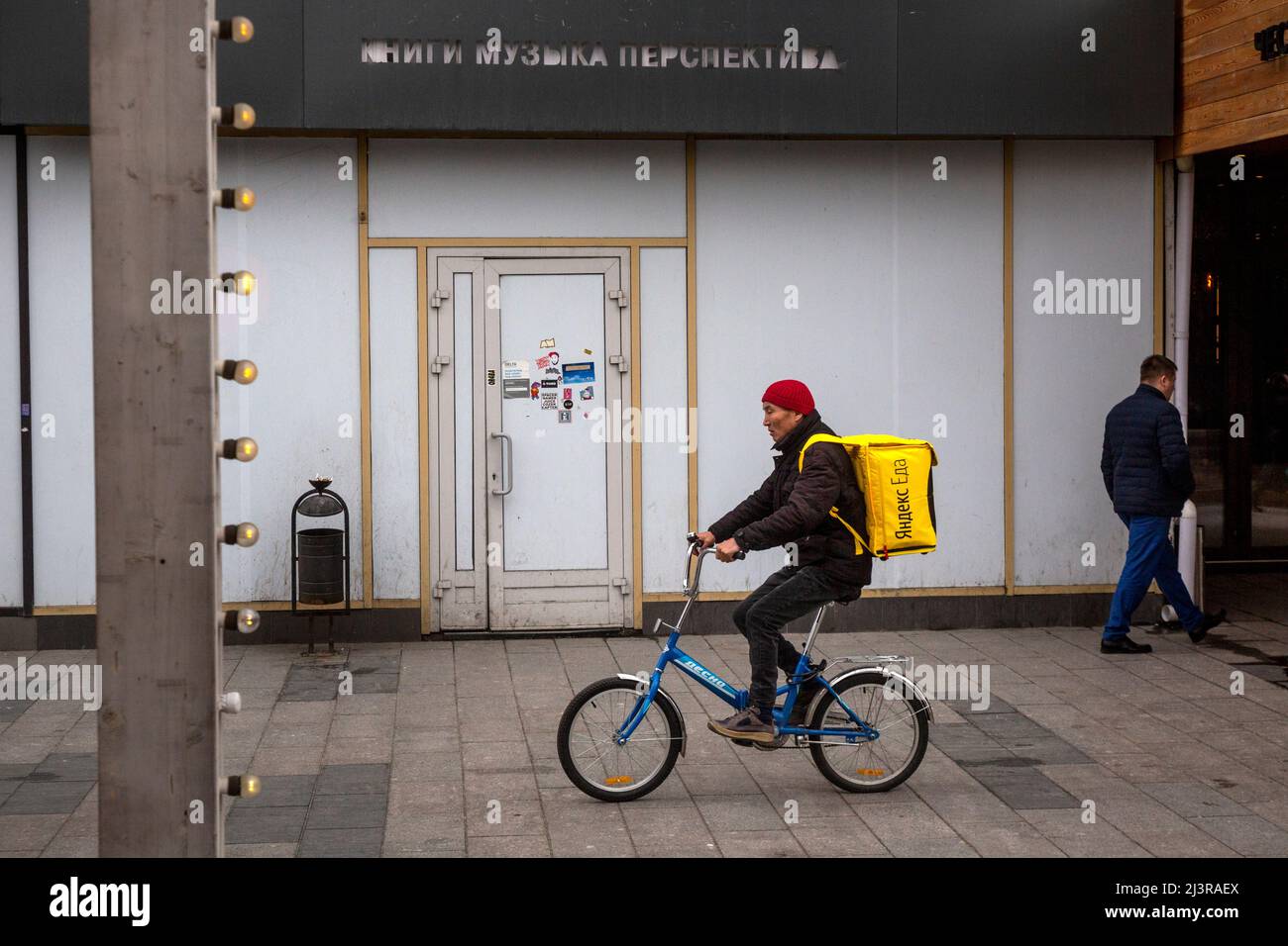 Moskau, Russland. 9.. April 2022. Ein Kurier des Yandex EDA-Zustelldienstes fährt mit dem Fahrrad entlang der Straße Novy Arbat vor dem Hintergrund eines geschlossenen Buchladens Respublika (eng: Republik) in Moskau, Russland. Die Inschrift über dem geschlossenen Eingang lautet „Perspektive der Buchmusik“. Stockfoto