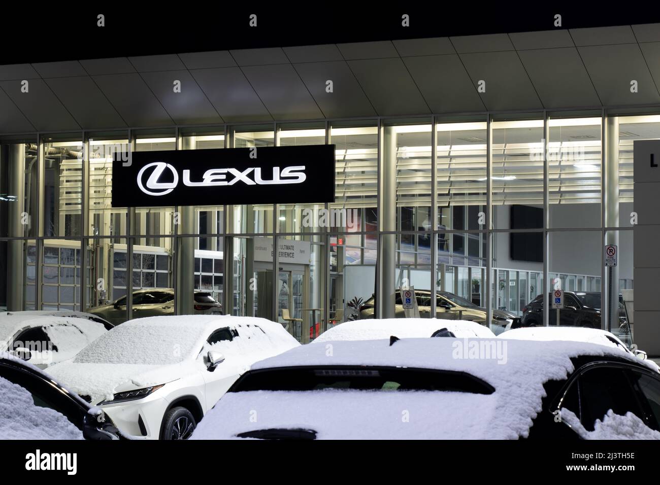Das Lexus-Logo auf einem hängenden Schild an der Vorderseite eines Lexus-Händlerbetriebs in der Nacht, von hinten gesehen von einem geparkten Neuwagen. Stockfoto