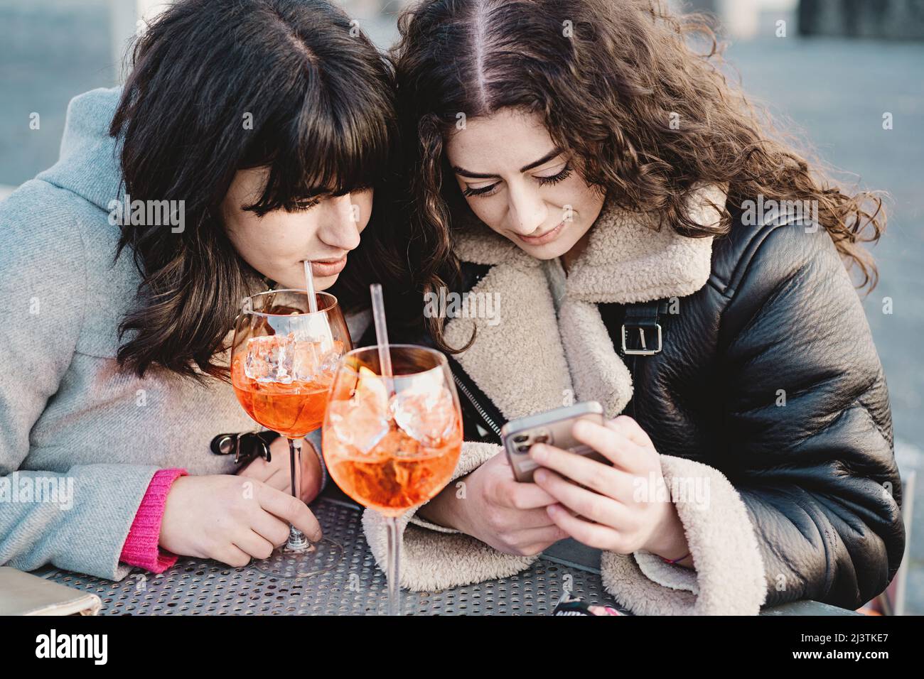 Zwei junge Frauen checken auf ihren Smartphones soziale Netzwerke, während sie zwei Spritz-Cocktails am Kaffeetisch trinken Stockfoto