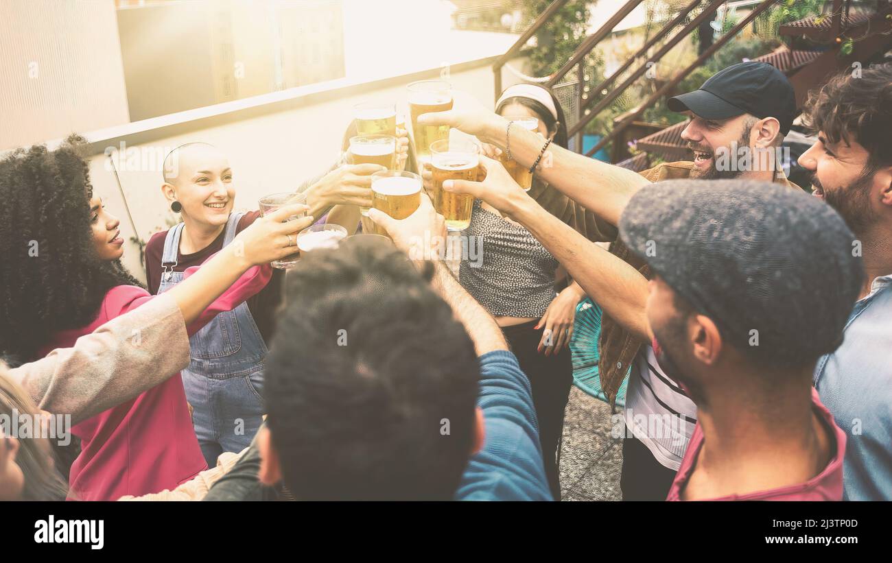 Fröhliche Gruppe von interracial Studenten mit Bier toasten. Fröhliche Menschen feiern auf dem Dach der Jugendherberge. Gefiltertes Bild mit Sonneneinstrahlung Stockfoto