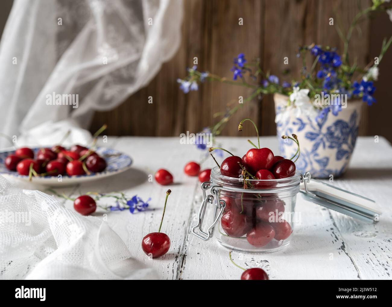 Frische Süßkirschen auf Teller und Glas auf weißem Holztisch mit blauen Blumen Stockfoto