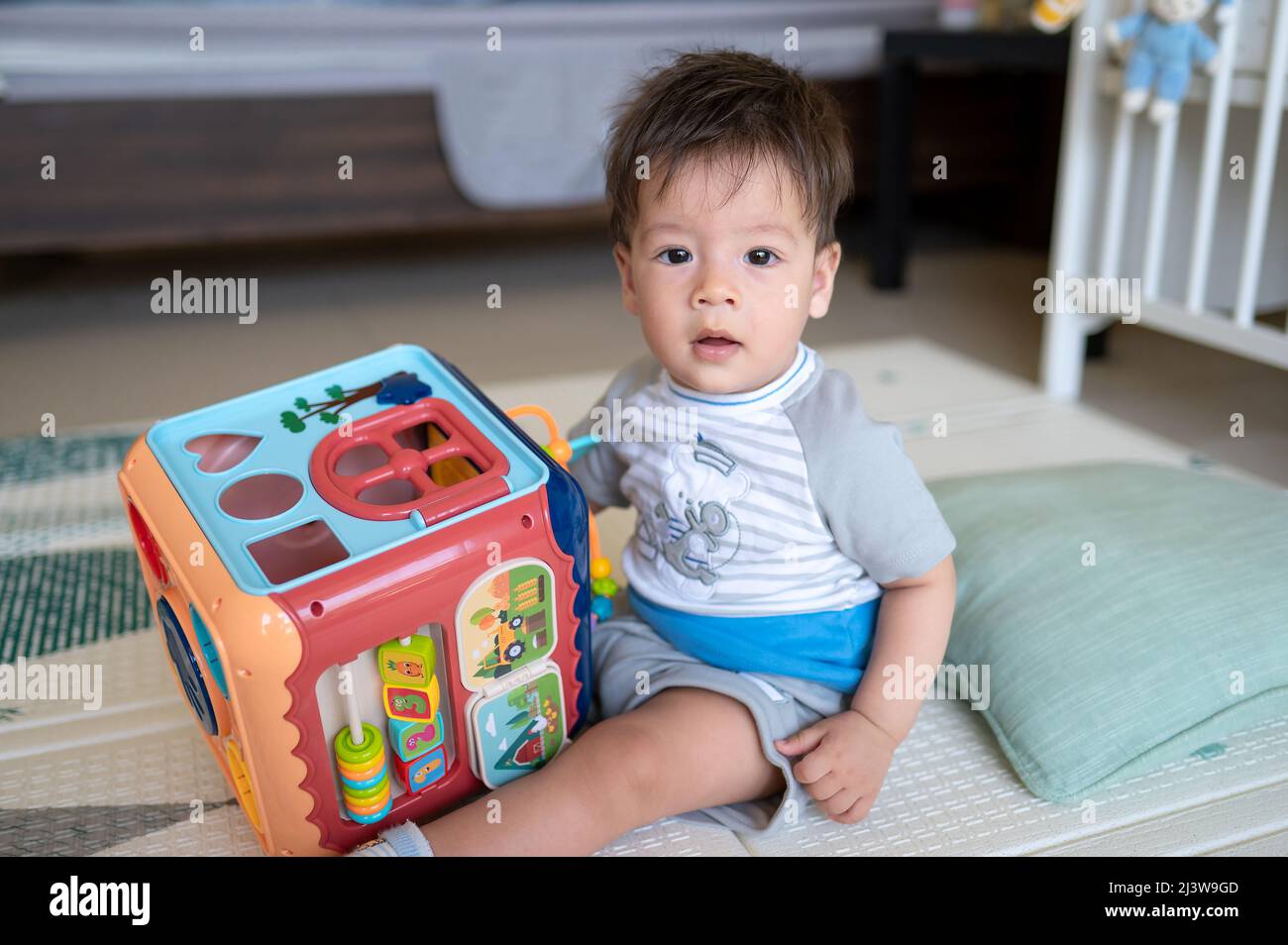 8 Monate alter Junge mit gemischter Rasse, der im Schlafzimmer mit einer Aktivitäts-Box spielt, während er auf dem Boden sitzt, der mit einer Gummispielmatte bedeckt ist Stockfoto