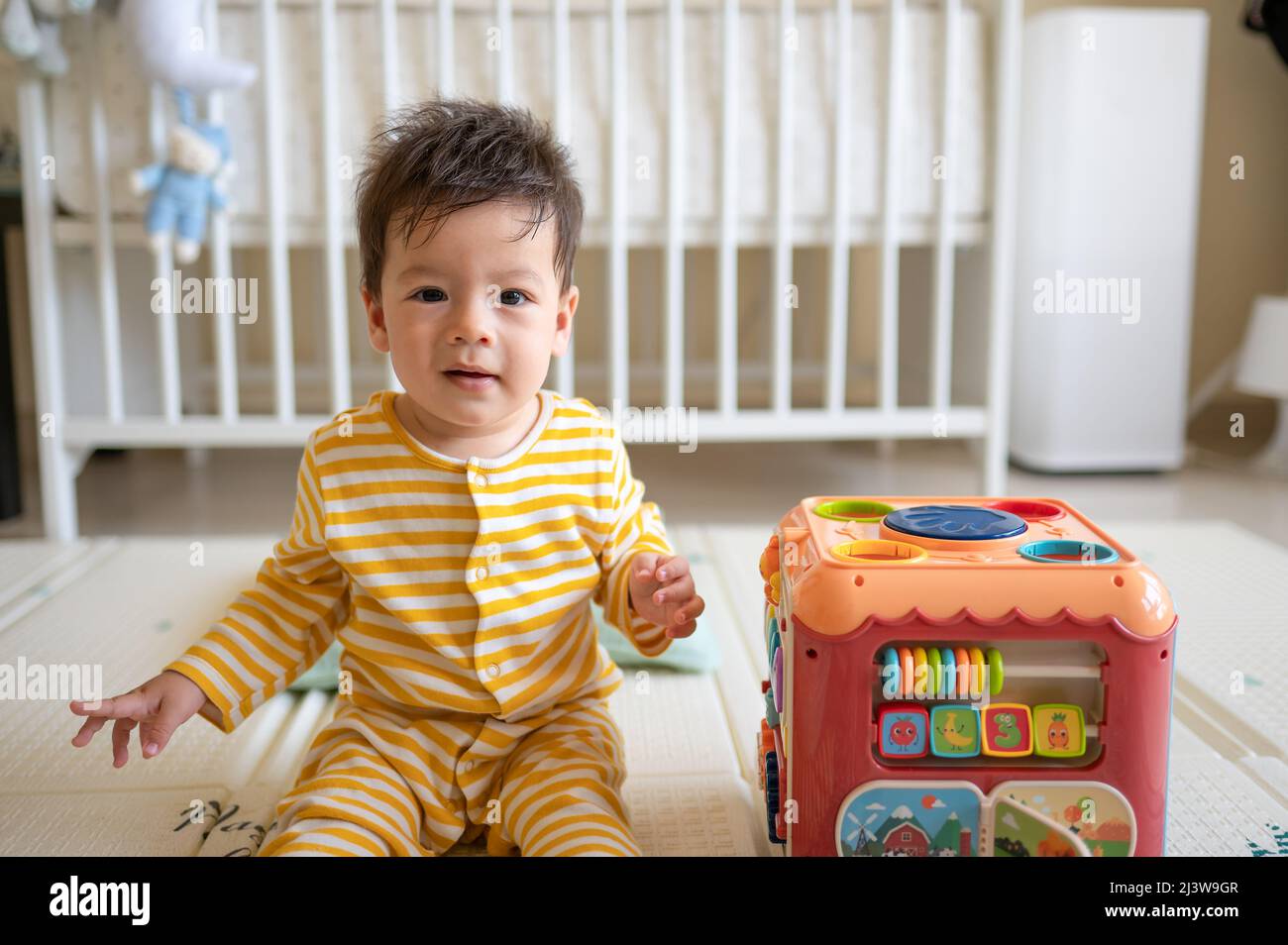 8 Monate alter Junge mit gemischter Rasse, der im Schlafzimmer mit einem gelben Pyjama auf dem Boden sitzt und mit einem Gummispiel bedeckt ist Stockfoto