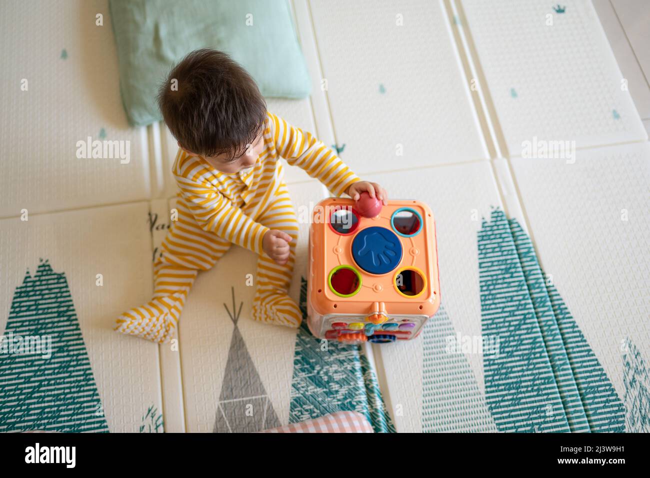 8 Monate alter Junge mit gemischter Rasse, der im Schlafzimmer mit einem gelben Pyjama auf dem Boden sitzt und mit einem Gummispiel bedeckt ist Stockfoto