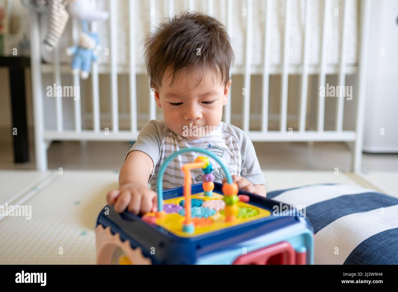 8 Monate alter Junge mit gemischter Rasse, der im Schlafzimmer mit einer Aktivitäts-Box spielt, während er auf dem Boden sitzt, der mit einer Gummispielmatte bedeckt ist Stockfoto