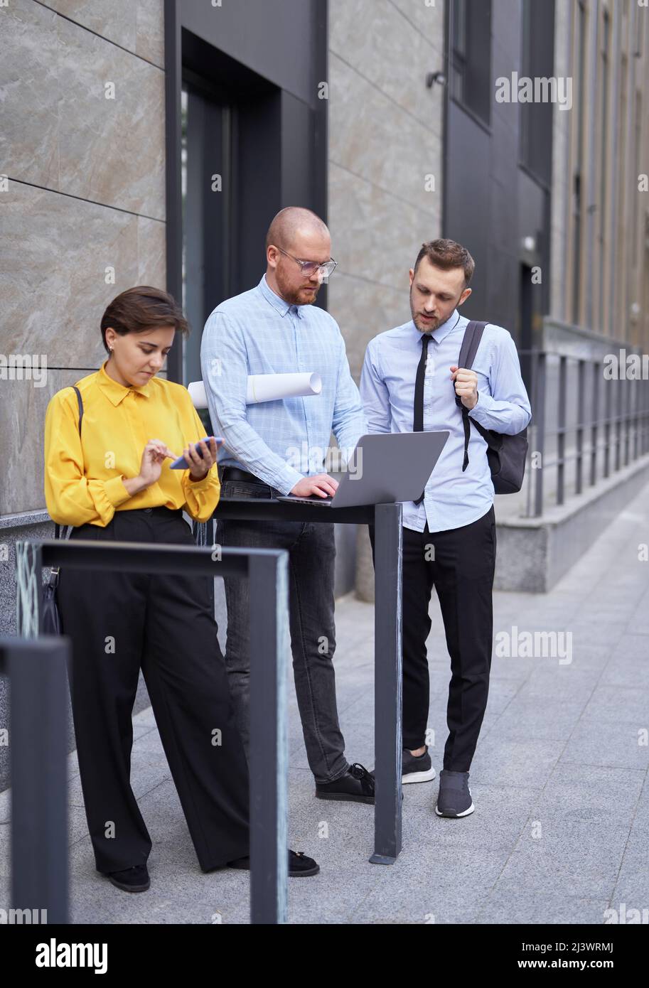 Geschäftsleute oder Team von Immobilienmaklern beobachten und diskutieren Projektpräsentation im Freien. Männer und Frauen in legerer Kleidung stehen draußen, benutzen Laptop, reden und Brainstorming Stockfoto