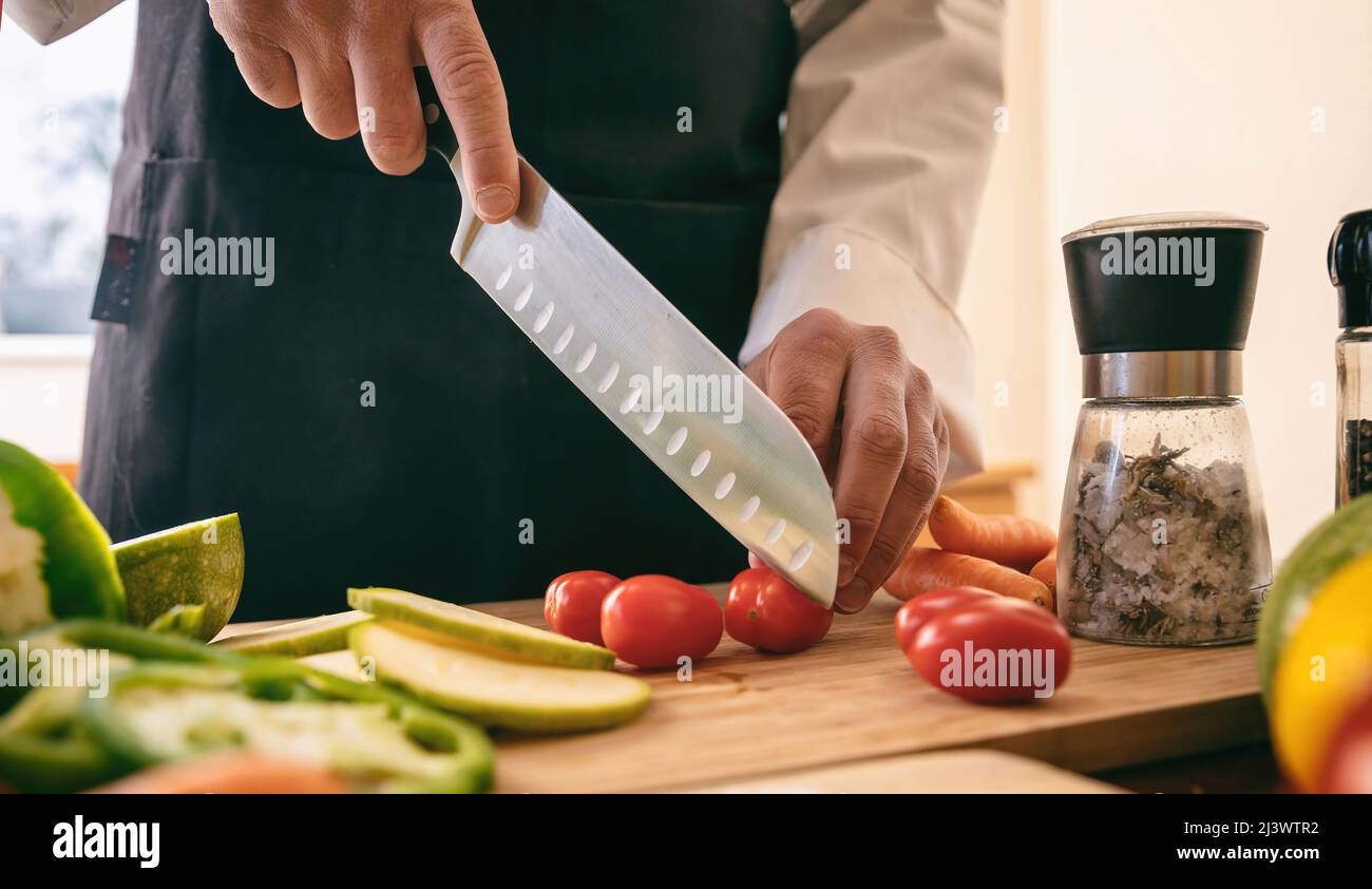 Koch mit schwarzer Schürze, die Tomate auf einem Schneidebrett schneidet, Hand kochen und ein Messer aus nächster Nähe halten. Frisches Gemüse auf Küchentisch, Restaurant vegetarisch Stockfoto