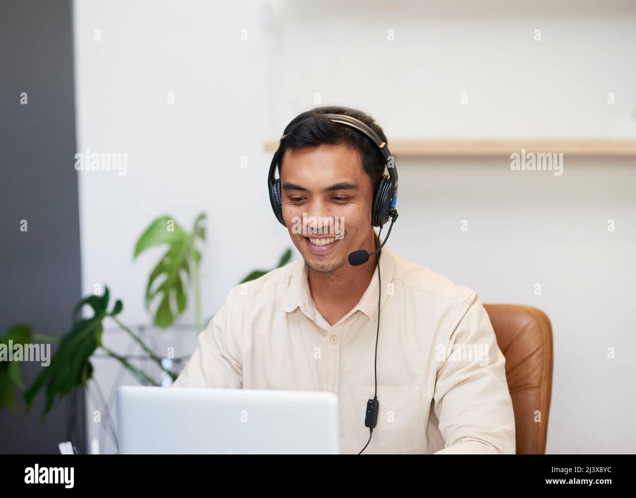 Ein asiatischer Mann lächelt, während er ein Headset trägt und im Büro auf seinen Laptop schaut Stockfoto