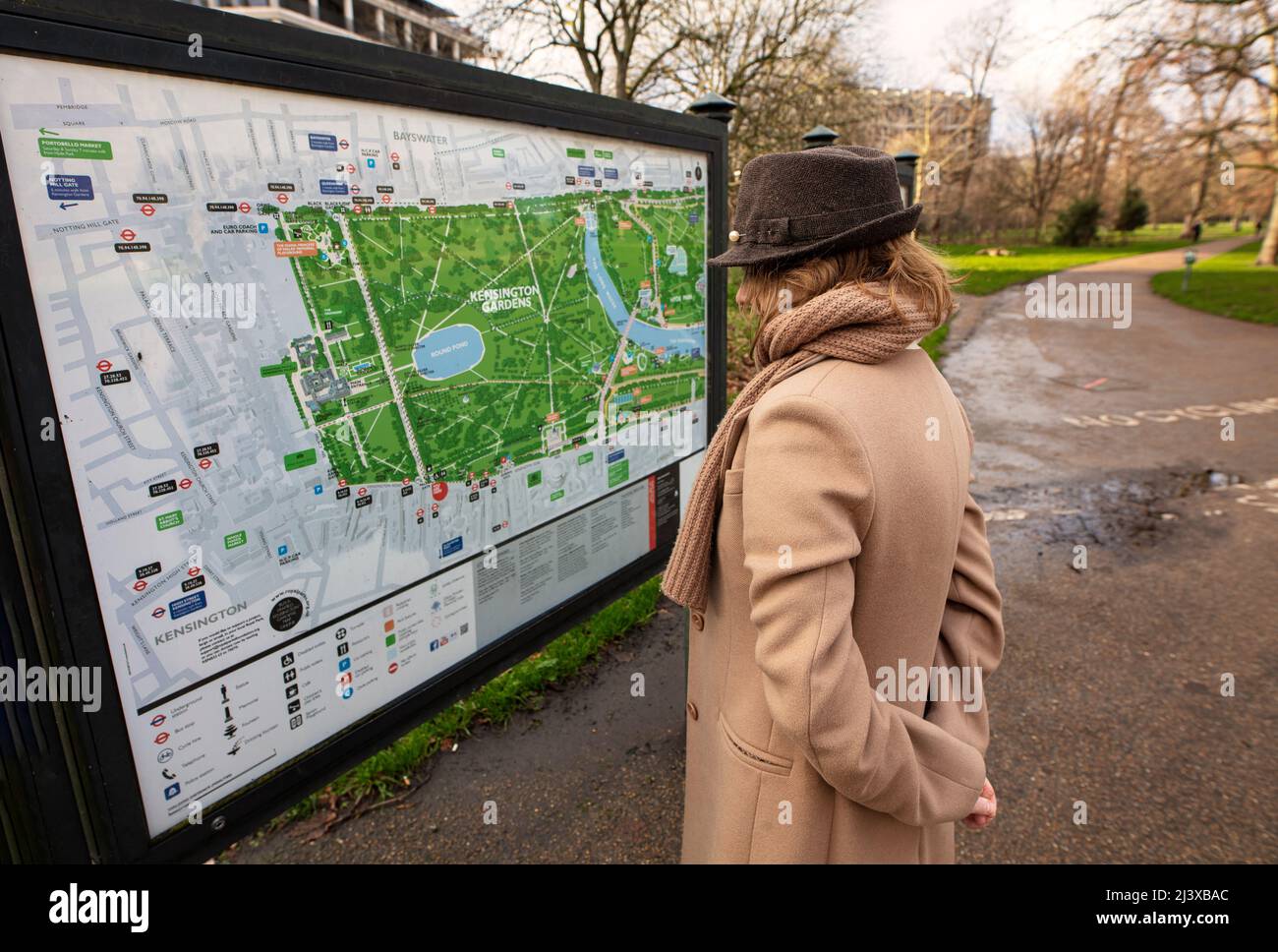 Frau mit Trilby-Hut, Schal und Kaschmirmantel, die das Schild im Eingang zu Kensington Gardens, London, untersucht Stockfoto