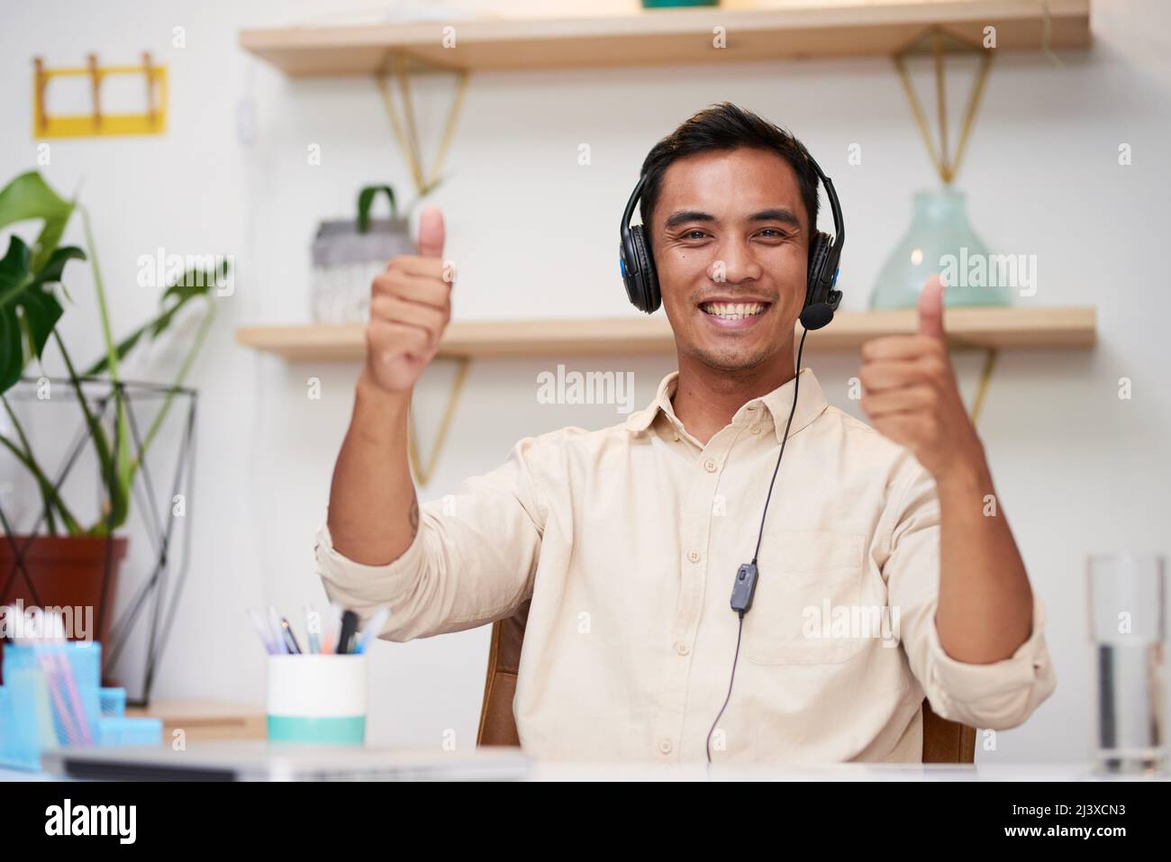 Ein junger Mann aus Asien hält die Daumen hoch, während er im Büro ein Callcenter-Headset trägt Stockfoto