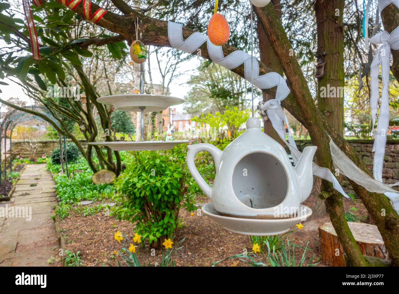 Eine Teekannen- und Kuchengarbe, die in einem öffentlichen Garten in Penrith, Cumbria, Großbritannien, an Baumzweigen hängen, ähneln der Teeparty von Mad Hatter Stockfoto