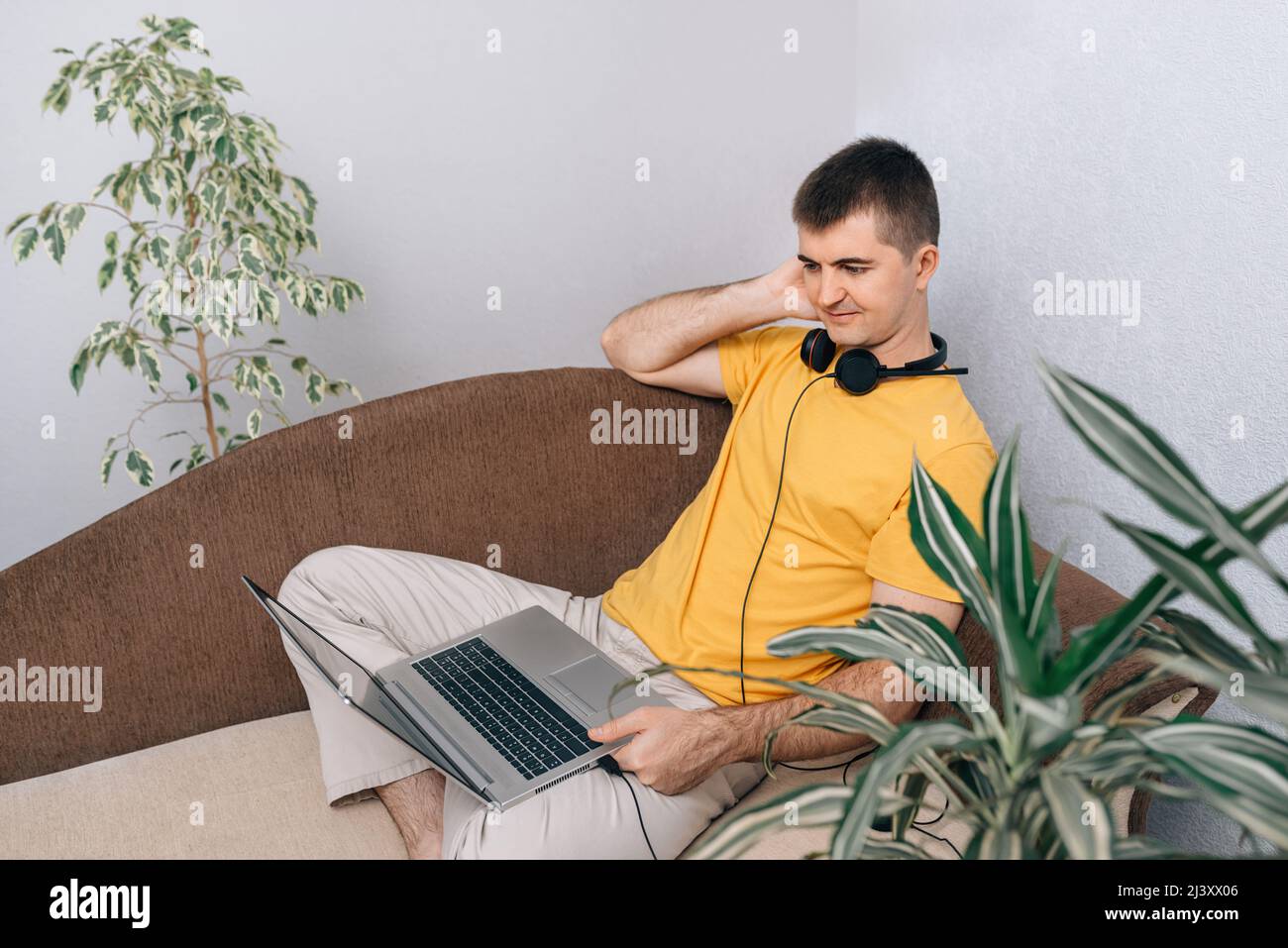 Ein junger lächelnder Mann in einem gelben T-Shirt arbeitet von zu Hause aus auf einem Laptop und kommuniziert online mit dem Kunden. Grüne Pflanzen im Zimmer. Konzept der Online-Bildung und Hausaufgaben, freiberuflich. Stockfoto