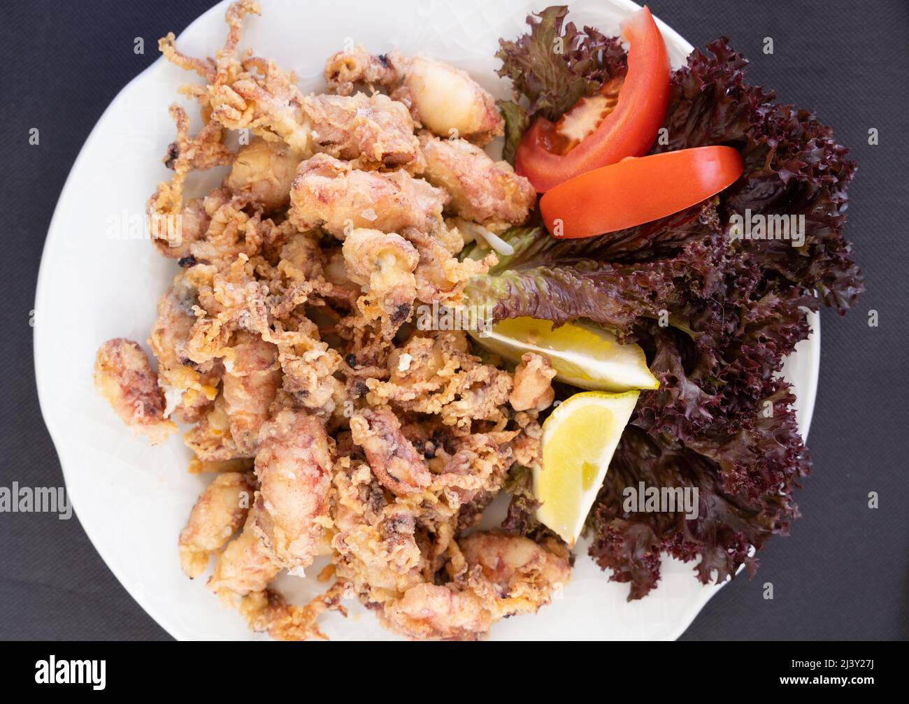 Draufsicht auf frittierte Baby-Tintenfische auf dem Teller mit Zitrone, Salat und Tomate. Traditionelle spanische Tapas mit Meeresfrüchten Stockfoto