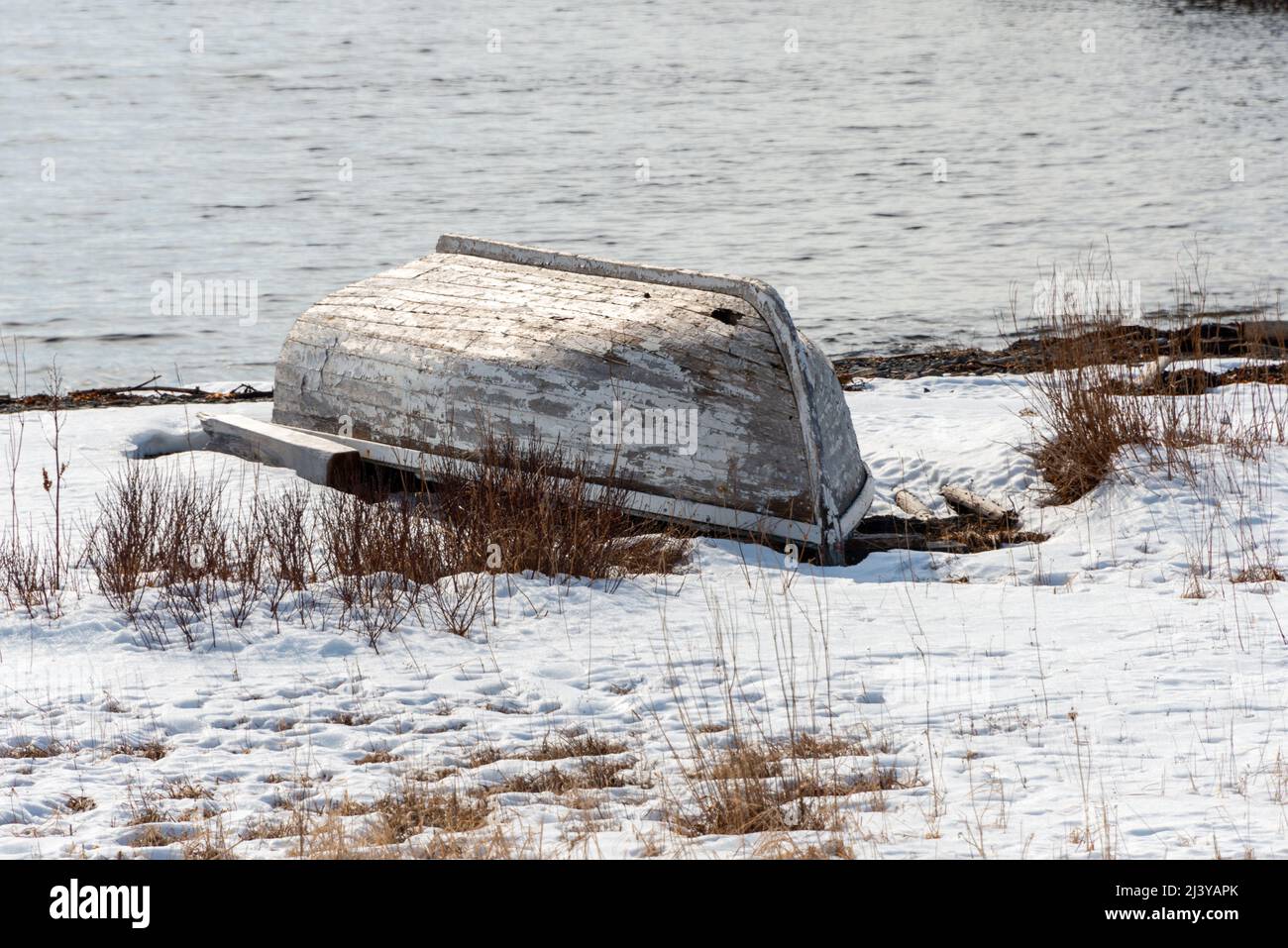 Ein weißes Fischerboot aus Holz mit abblätternder Farbe, das neben dem Meer und der felsigen Küste mit Schnee auf dem Boden befahren wurde. Das offene Boot ist von unten nach oben. Stockfoto