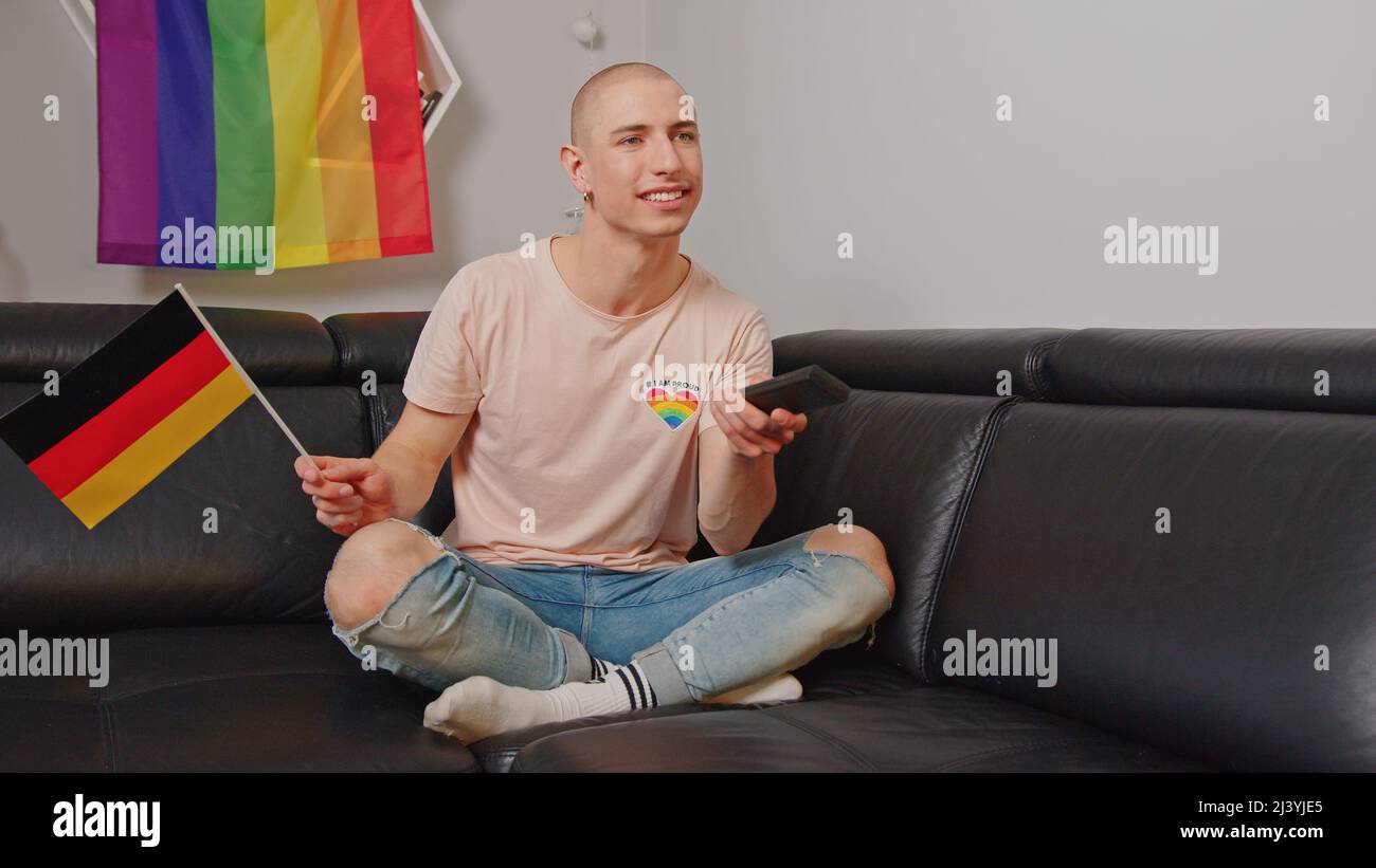 Stolzer schwuler europäischer Influencer, der in Deutschland lebt und die deutsche Flagge schwenkt, während er auf einem Sofa sitzt und die Wahlen beobachtet. Hochwertige Fotos Stockfoto