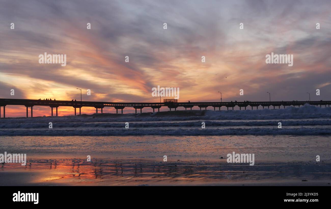 Silhouette von Menschen, die spazieren gehen, Surfer, die am Pier im Meerwasser surfen. Meereswellen, dramatischer Himmel bei Sonnenuntergang. Kalifornische Küste, Strand oder Ufer bei Sonnenuntergang. Sommer Seesaat nahtlose Schleife Cinemagraph. Stockfoto