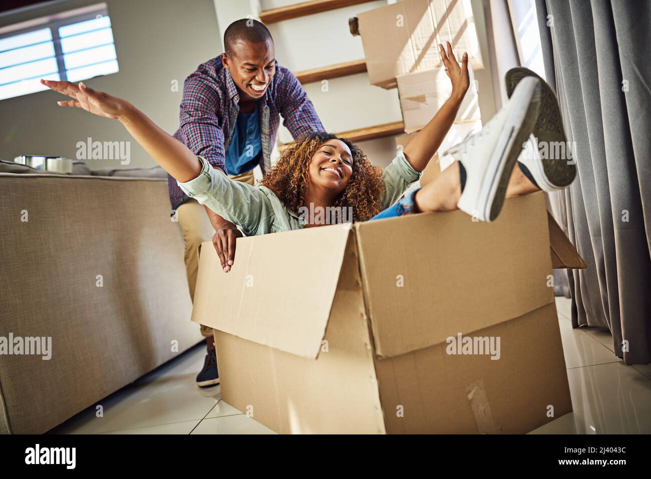 Ich werde Sie in seiner Schachtel schicken. Aufnahme einer fröhlichen jungen Frau in einer Kiste mit ihrem Partner, der die Kiste tagsüber zu Hause nach innen drückt. Stockfoto