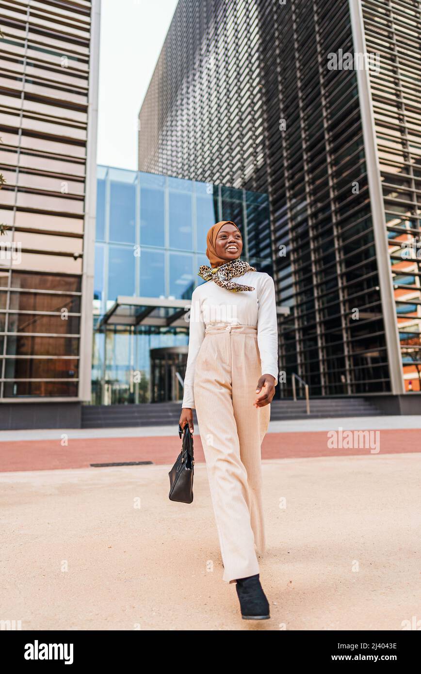 Ganzer Körper der glücklichen afroamerikanischen Frau in elegantem Casual-Outfit und traditionellem Kopftuch, die mit einer Tasche im zentralen Bezirk gegen den hohen Turm läuft und mit einem breiten Lächeln wegschaut Stockfoto