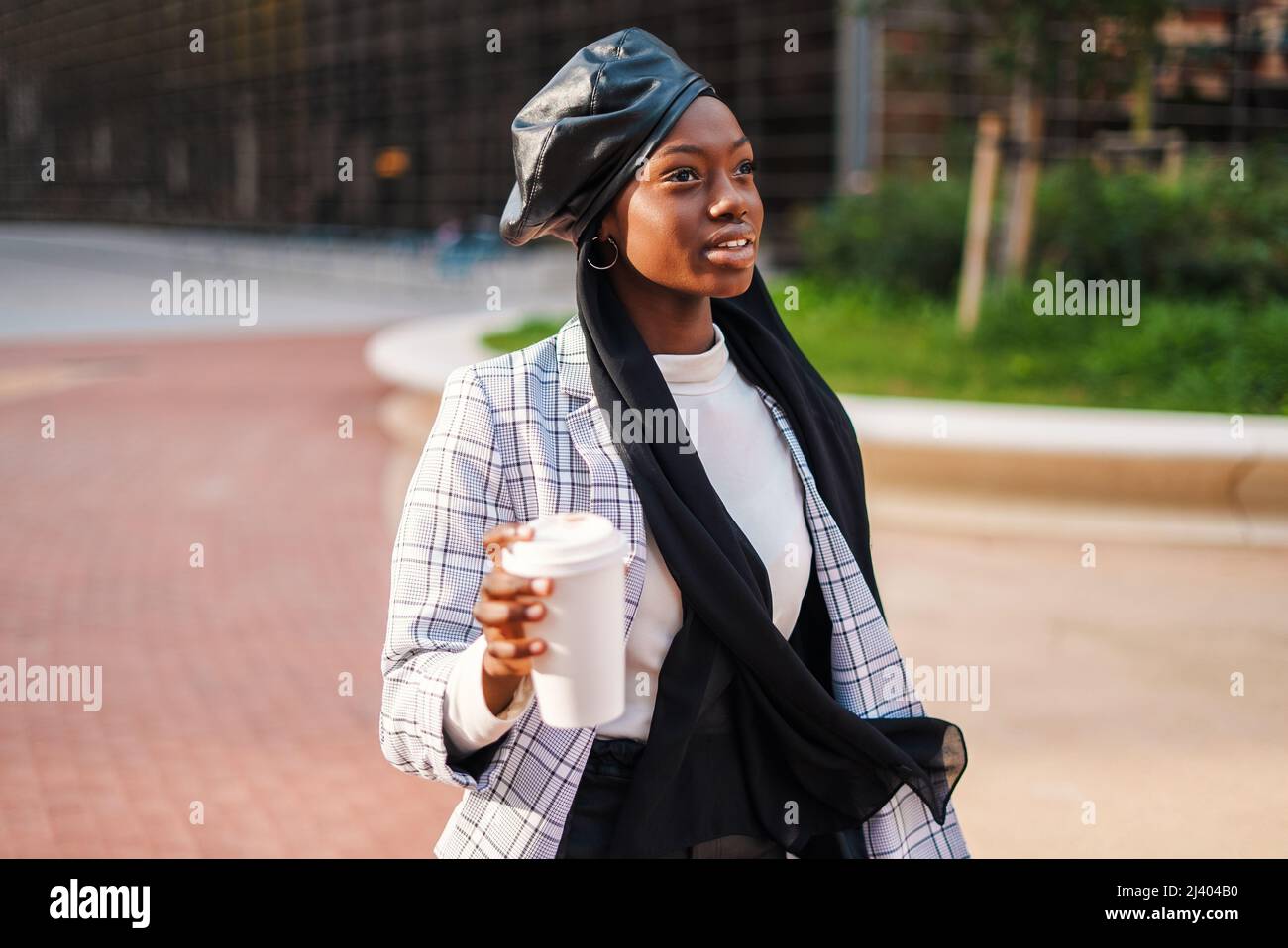 Seriöse junge afroamerikanische Dame in trendigem Outfit mit Lederberette auf muslimischem Kopftuch, mit einem Getränk zum Mitnehmen und mit aufmerksamem Blick im Park Stockfoto