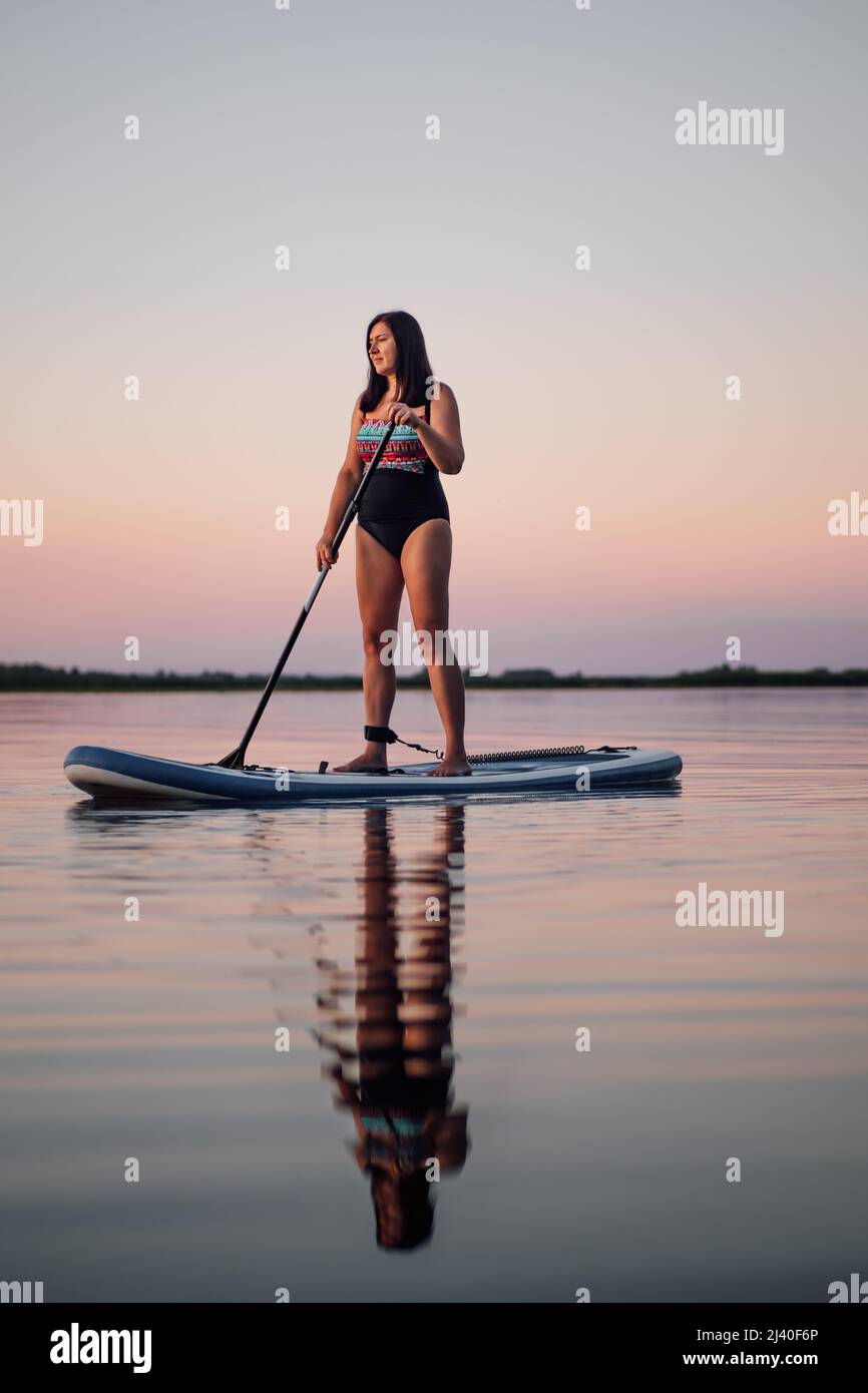 Porträt einer ruhigen, entspannten Frau mittleren Alters, die mit Ruder in den Händen auf den See blickt, mit Spiegelung im Wasser und rosa Himmel im Hintergrund Stockfoto