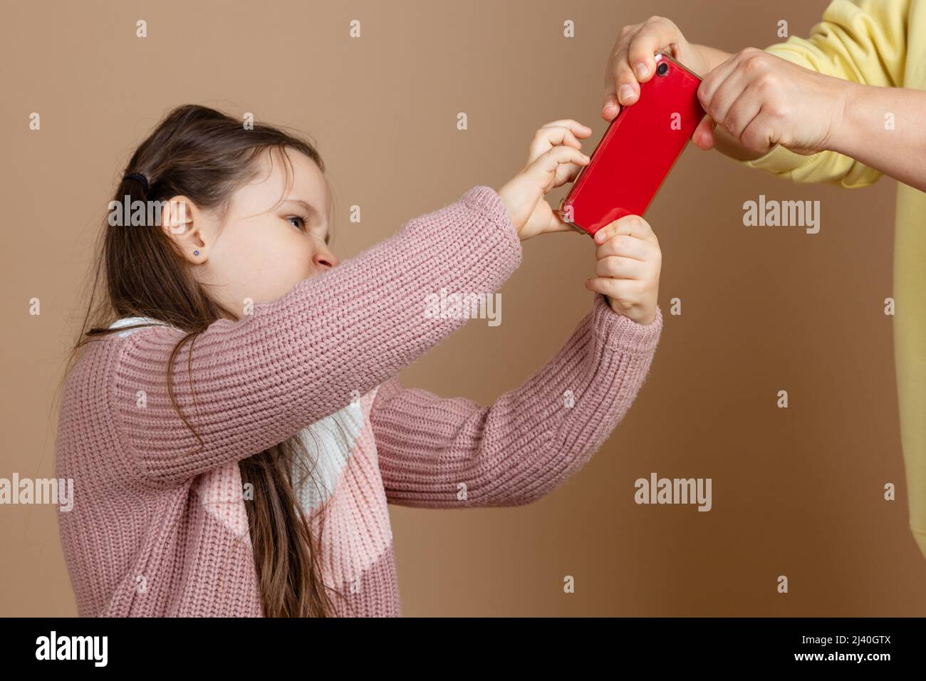 Portrait der Eltern, die das Smartphone aus den Händen der Mädchen nehmen, beige Hintergrund. Verärgerte Tochter hält das Telefon fest und widersetzt sich. Konzept der Prävention von Stockfoto