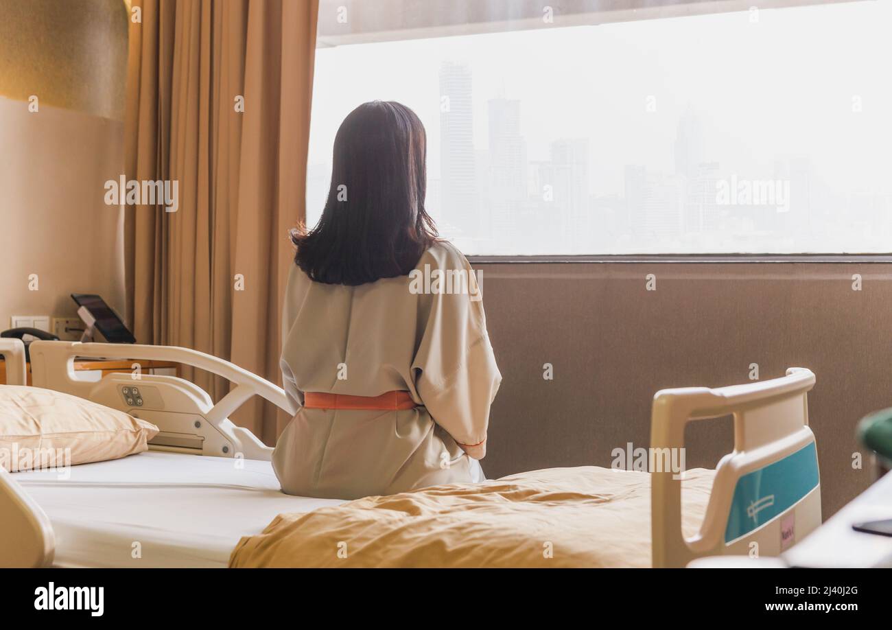 Rückansicht einer geduldigen Frau, die auf der Krankenstation auf dem Bett saß und auf das Fenster schaute. Stockfoto