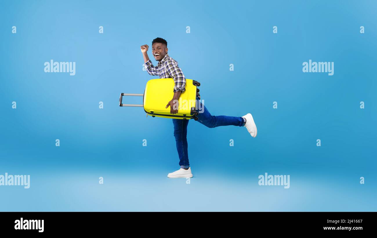 Voller Länge aufgeregt schwarzen Kerl mit Koffer zum Flughafen laufen, bereit, Sommerurlaub auf blauen Studio-Hintergrund zu beginnen Stockfoto