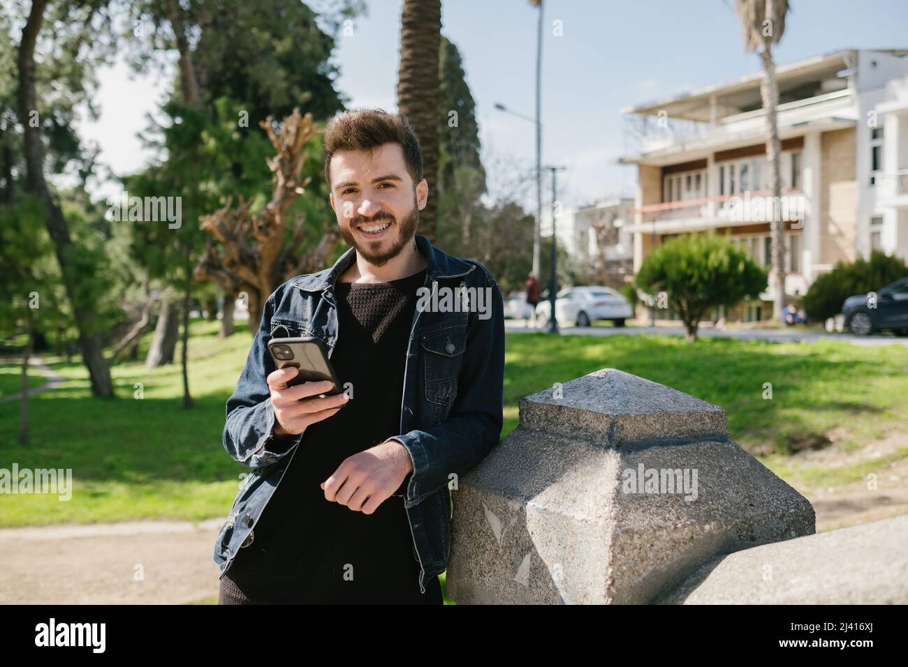 Junger Mann mit blauer jean-Jacke, der sich in einem Park an einen dekorativen Stein lehnt. Blick auf die Kamera, während Sie ein Telefon beim Scrollen, Messaging oder Ansehen von V halten Stockfoto