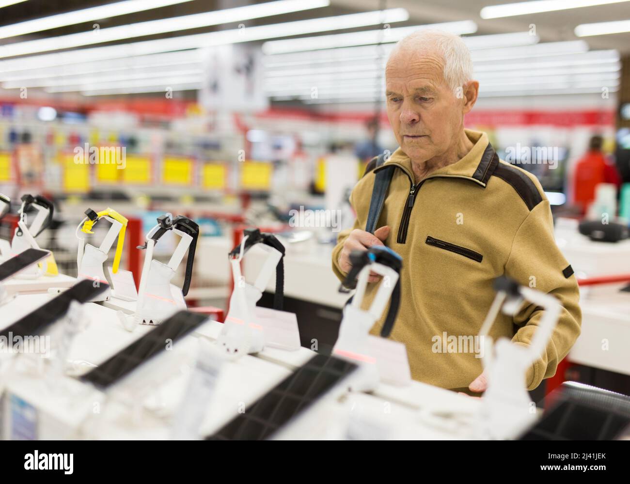 Älterer grauhaariger Rentner, der im Ausstellungsraum des digitalen Warenhauses einen Schalter mit elektronischen Geräten und intelligenten Uhren untersucht Stockfoto