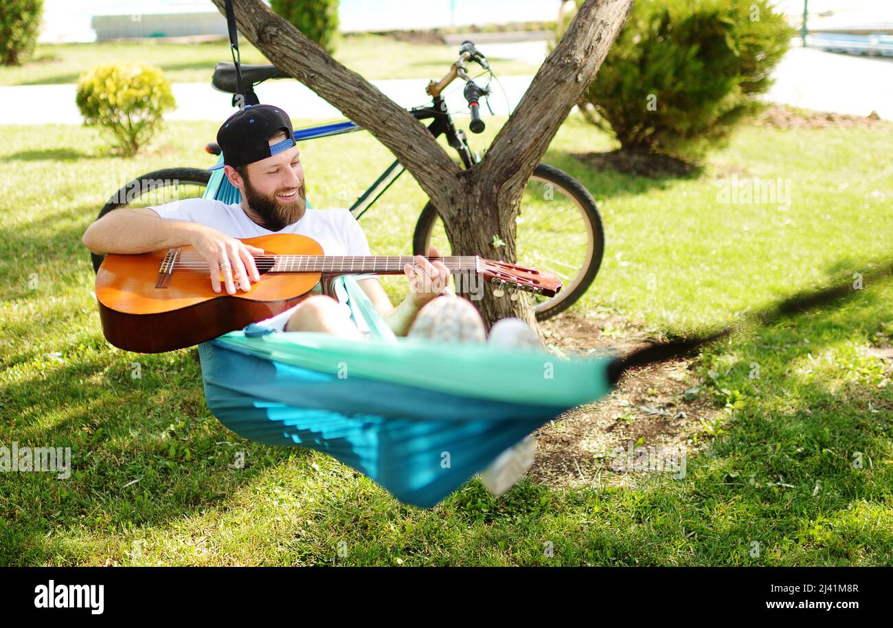 Bärtiger Mann mit einer Gitarre, der in einer Hängematte liegt und vor dem Hintergrund von Natur und Grün lächelt. Stockfoto