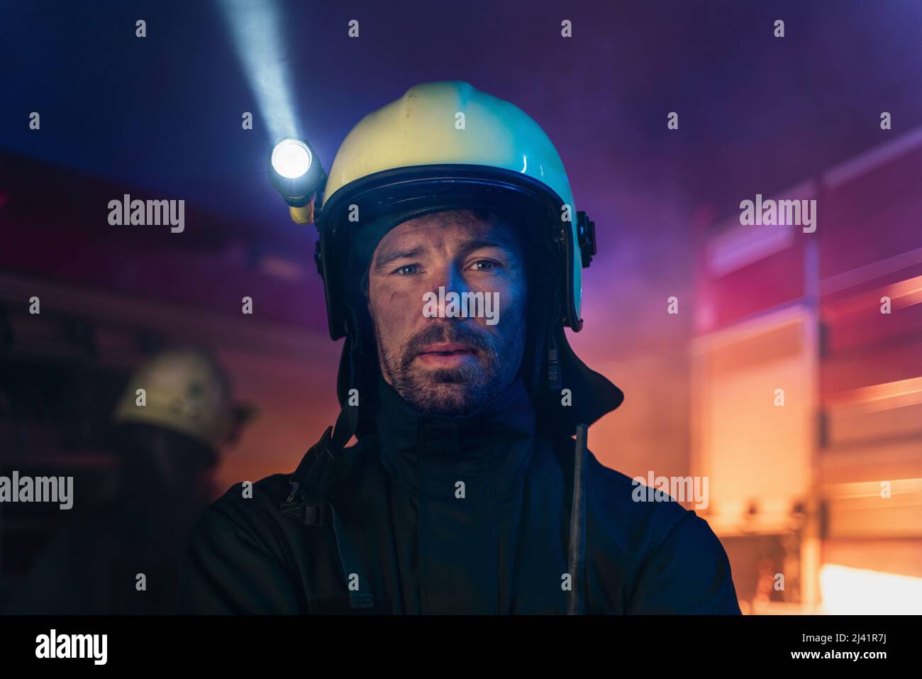 Porträt eines schmutzigen Feuerwehrmanns im Dienst mit Feuerwehrauto im Hintergrund in der Nacht, Blick auf die Kamera. Stockfoto