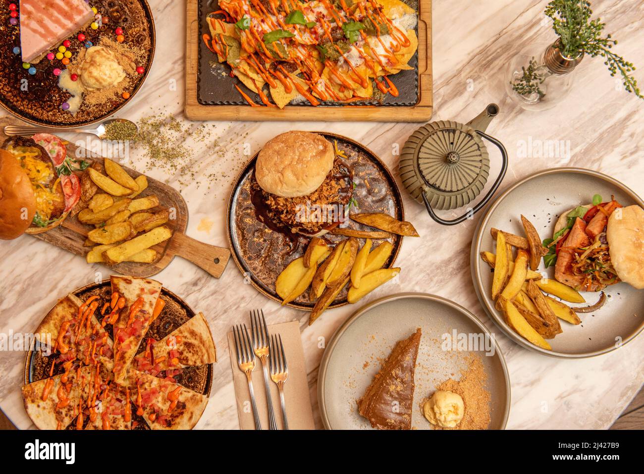 Set aus hausgemachten Fast-Food-Gerichten mit verschiedenen Burgern, Nachos mit Guacamole und Nacho-Käse, hausgemachten Pommes frites, verschiedenen Kuchen auf einem Marmortisch Stockfoto