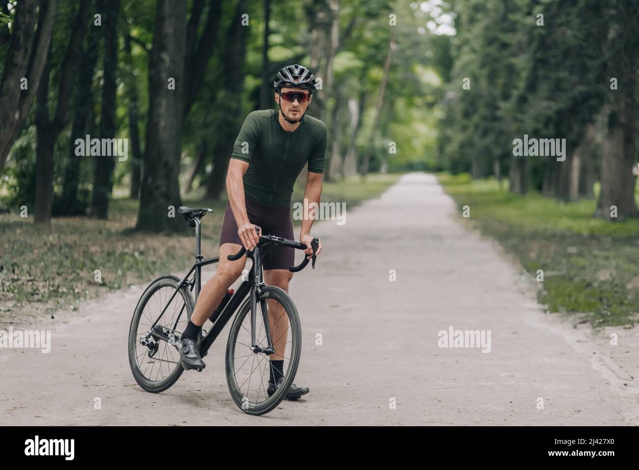 Radprofi in Sportkleidung, Helm und Brille, die nach der Fahrt im grünen Park entspannen. Kaukasischer Mann sitzt auf schwarzem Fahrrad und Wiederherstellung der Leistung f Stockfoto