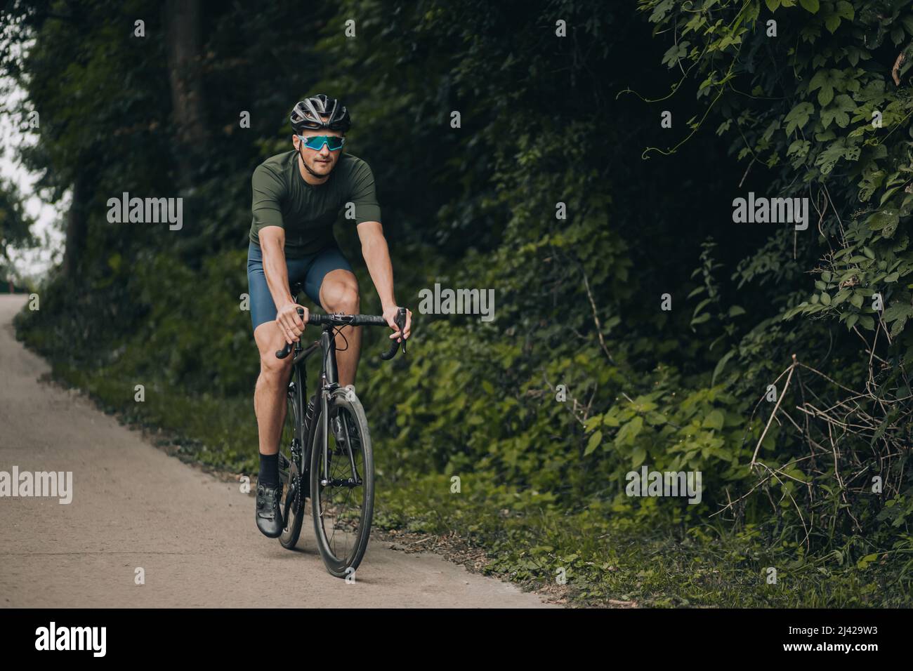 Muskulöser kaukasischer Mann in Sportkleidung, Sicherheitsbrille und Spiegelbrille, der im grünen Park Fahrrad fährt. Gesund und stark Kerl verbringen Freizeit für Stockfoto