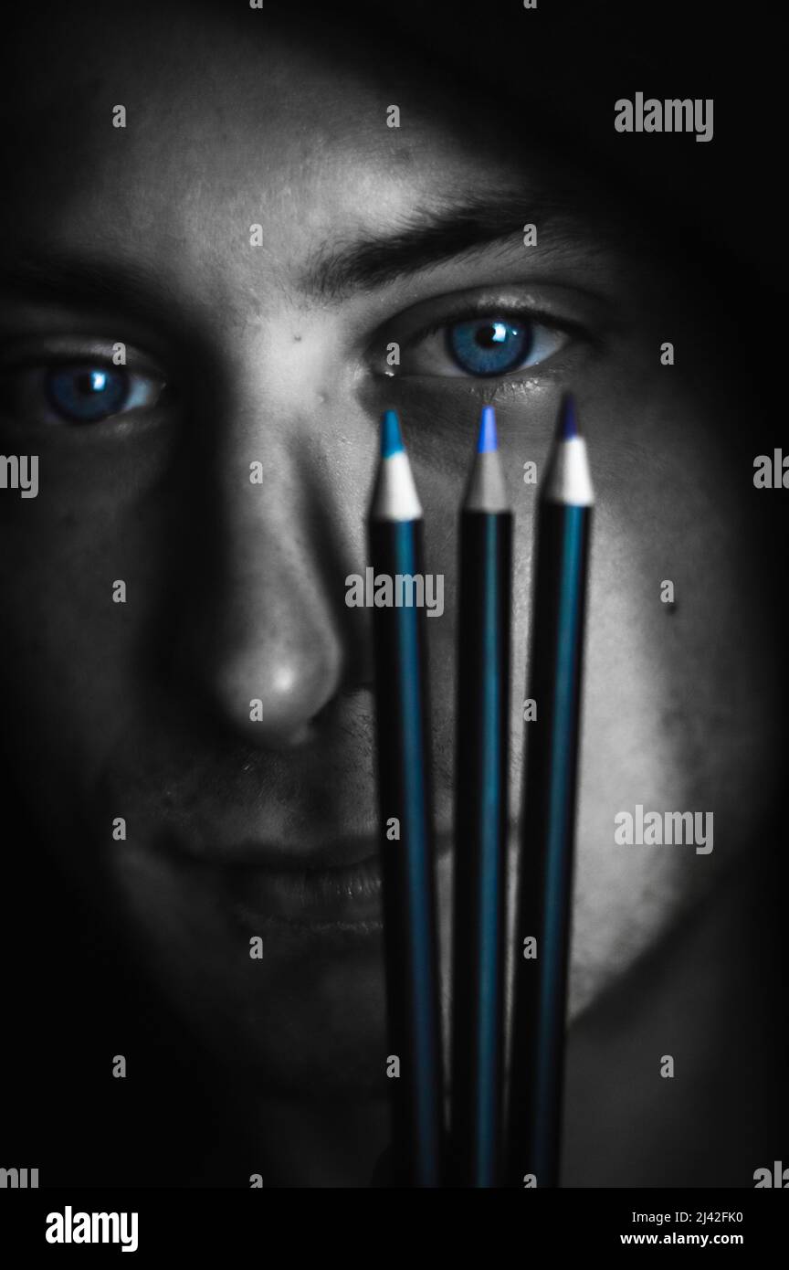 Passende Farben - Schwarz-Weiß-Porträt eines jungen blauäugigen Mannes, der drei Buntstifte in der Nähe seines Auges hält, um den richtigen Farbton zu finden Stockfoto