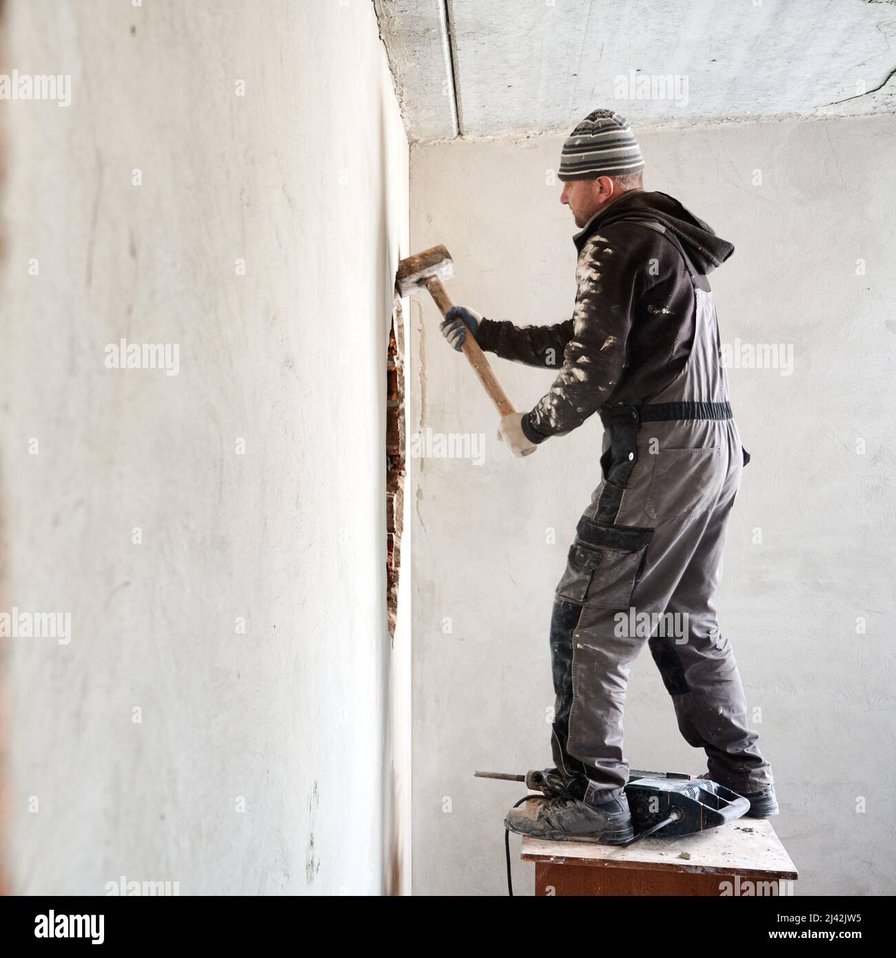 Erfahrener Arbeiter, der die Innenwand demontiert. Mann, der auf einer erhöhten Fläche steht, wo sich der Perforator befindet, und mit Hilfe eines Vorschlaghammers, der durch ein Loch in der Wand vergrößert wird. Stockfoto