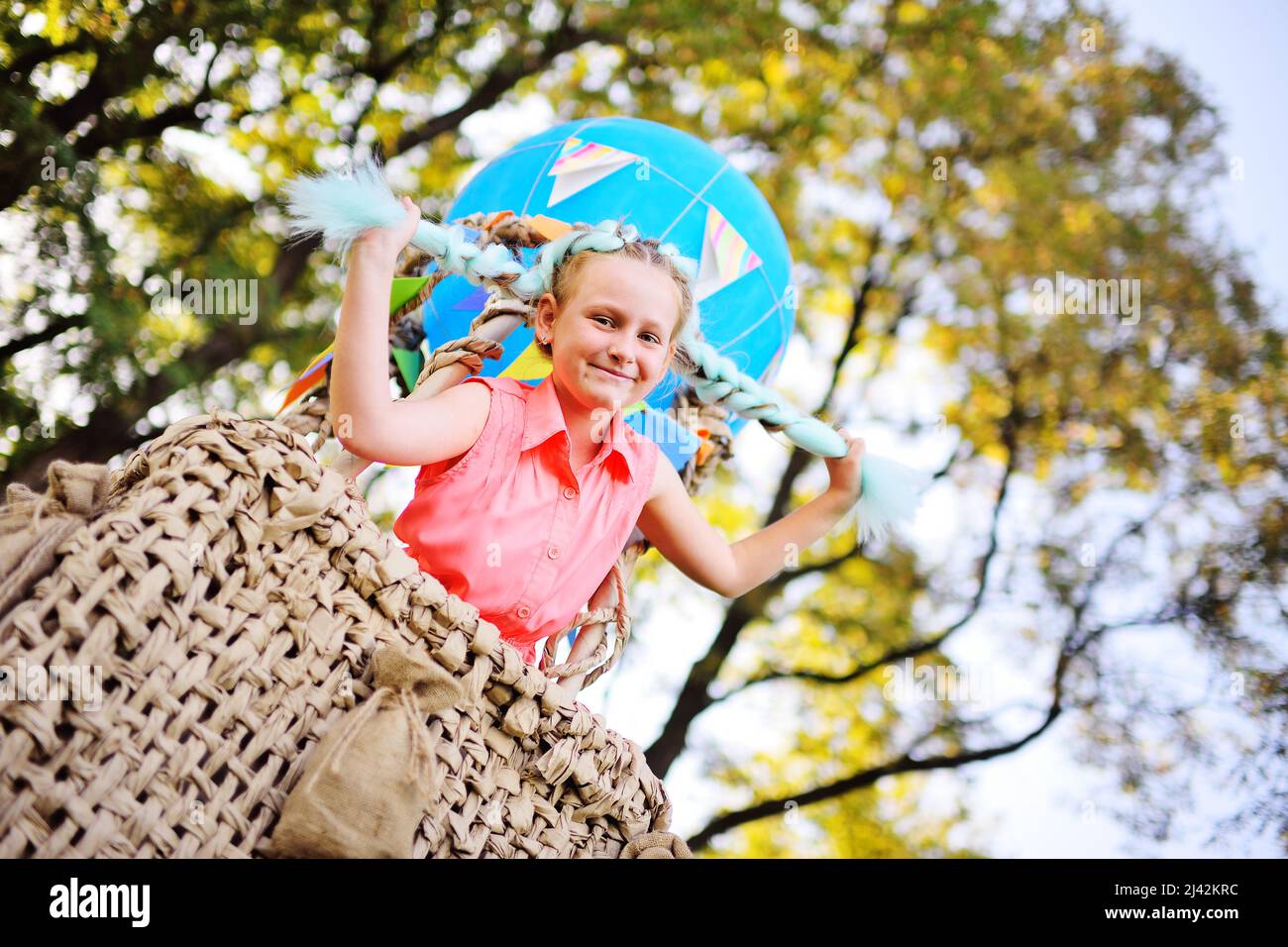 Kleines Mädchen in rosa Kleidung lächelt sitzen in einem Korb mit einem blauen Ballon vor einem Hintergrund von Sonnenlicht und Grün. Stockfoto