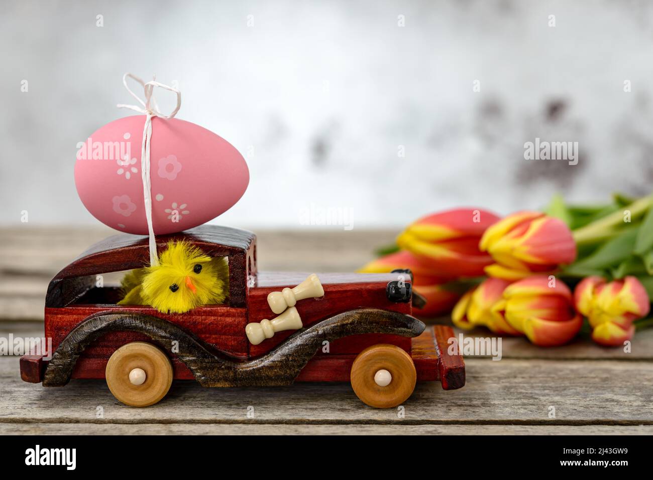 Ein Spielzeugauto aus Holz mit einem Osterei auf dem Dach und einem Hühnerfahrer neben einem Tulpenstrauß auf einem rustikalen Tisch. Stockfoto