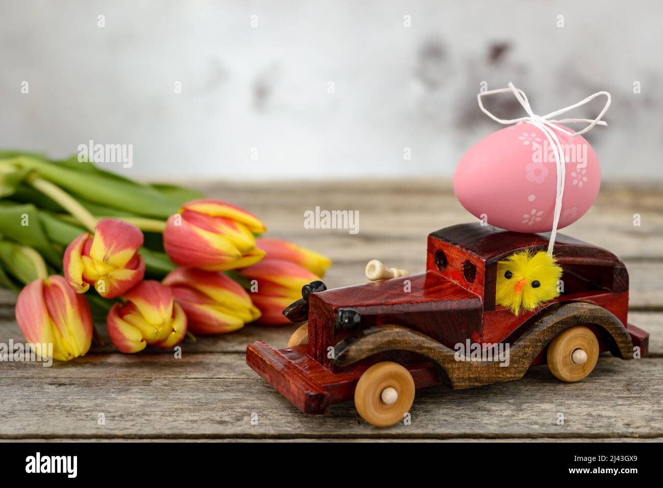 Ein Spielzeugauto aus Holz mit einem Osterei auf dem Dach und einem Hühnerfahrer neben einem Tulpenstrauß auf einem rustikalen Tisch. Stockfoto
