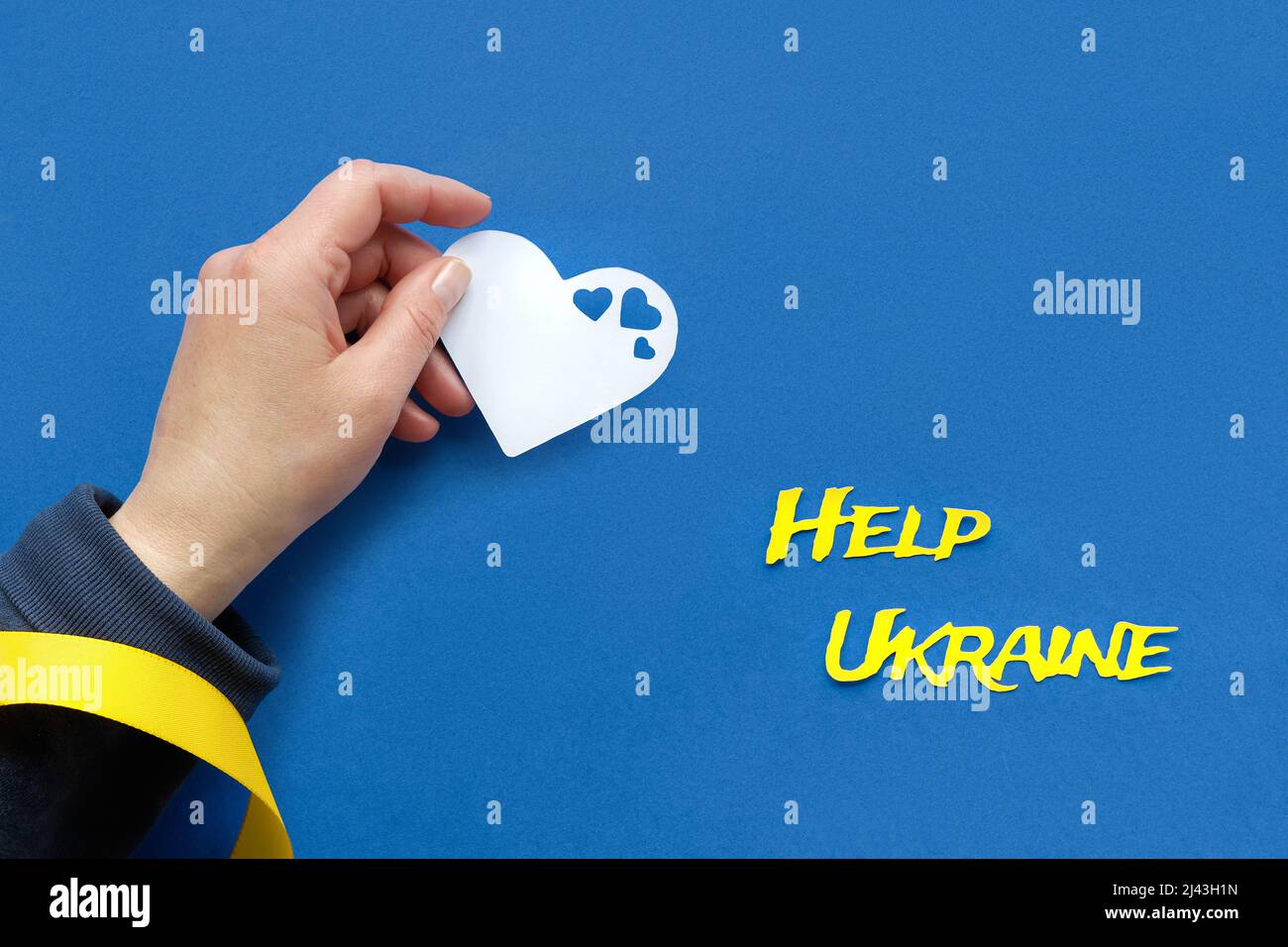 Text Hilfe Ukraine, Hand mit gelbem Band hält Herz Papier Silhouette auf blauem Hintergrund Stockfoto