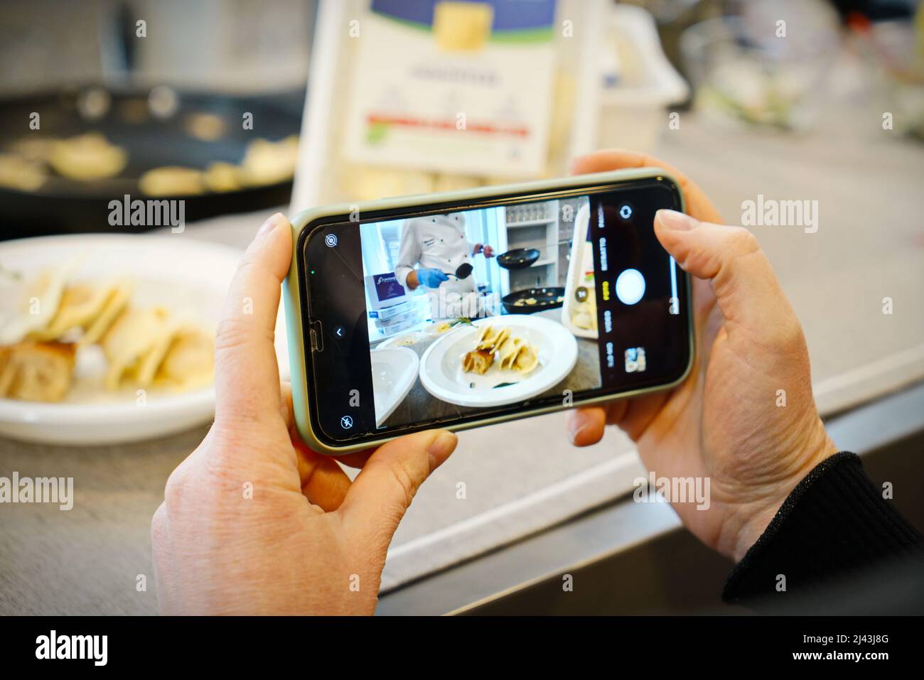 Hände, die Fotos mit dem Mobiltelefon von Koch in einer Küchenwerkstatt machen. Selektiver Fokus auf das Mobiltelefon im Vordergrund. Mailand, Italien - März 2022 Stockfoto