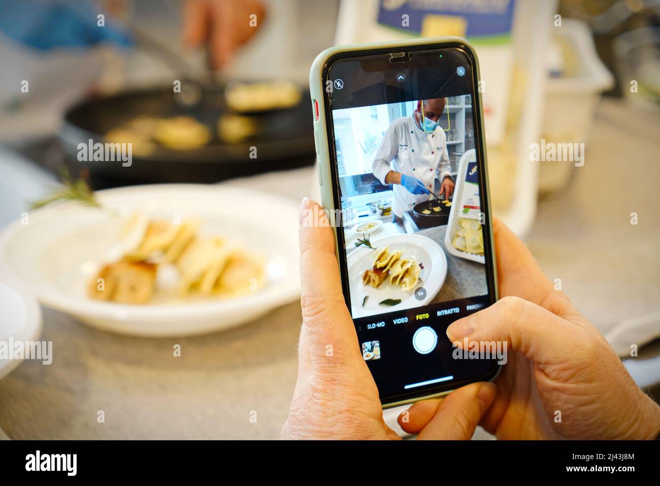 Hände, die Fotos mit dem Mobiltelefon von Koch in einer Küchenwerkstatt machen. Selektiver Fokus auf das Mobiltelefon im Vordergrund. Mailand, Italien - März 2022 Stockfoto