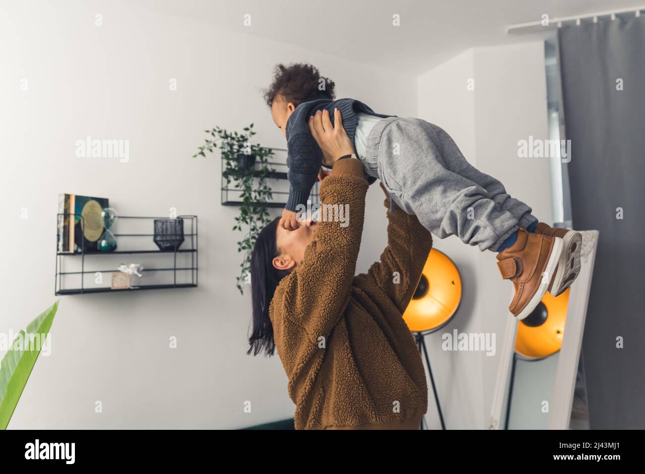 Eine schwarzhaarige Frau mit einem braunen Pullover, der einen gewellten kleinen Jungen im Zimmer mit weißen Zimmern mit mittlerer Schussaufnahme nach oben hält. Hochwertige Fotos Stockfoto