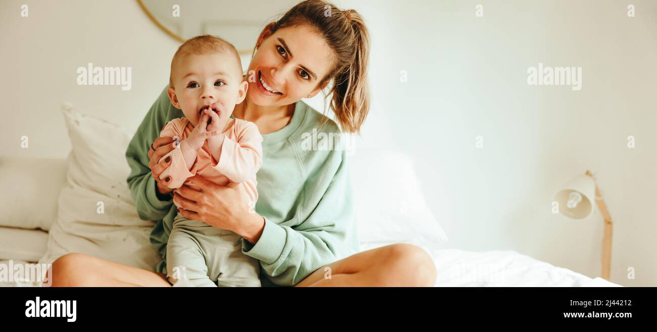 Fröhliche Mutter lächelt an der Kamera, während sie ihr entzückendes Baby hält. Glückliche junge Mutter, die mit ihrem neugeborenen Baby auf dem Bett sitzt. Liebende Mutter Kreati Stockfoto