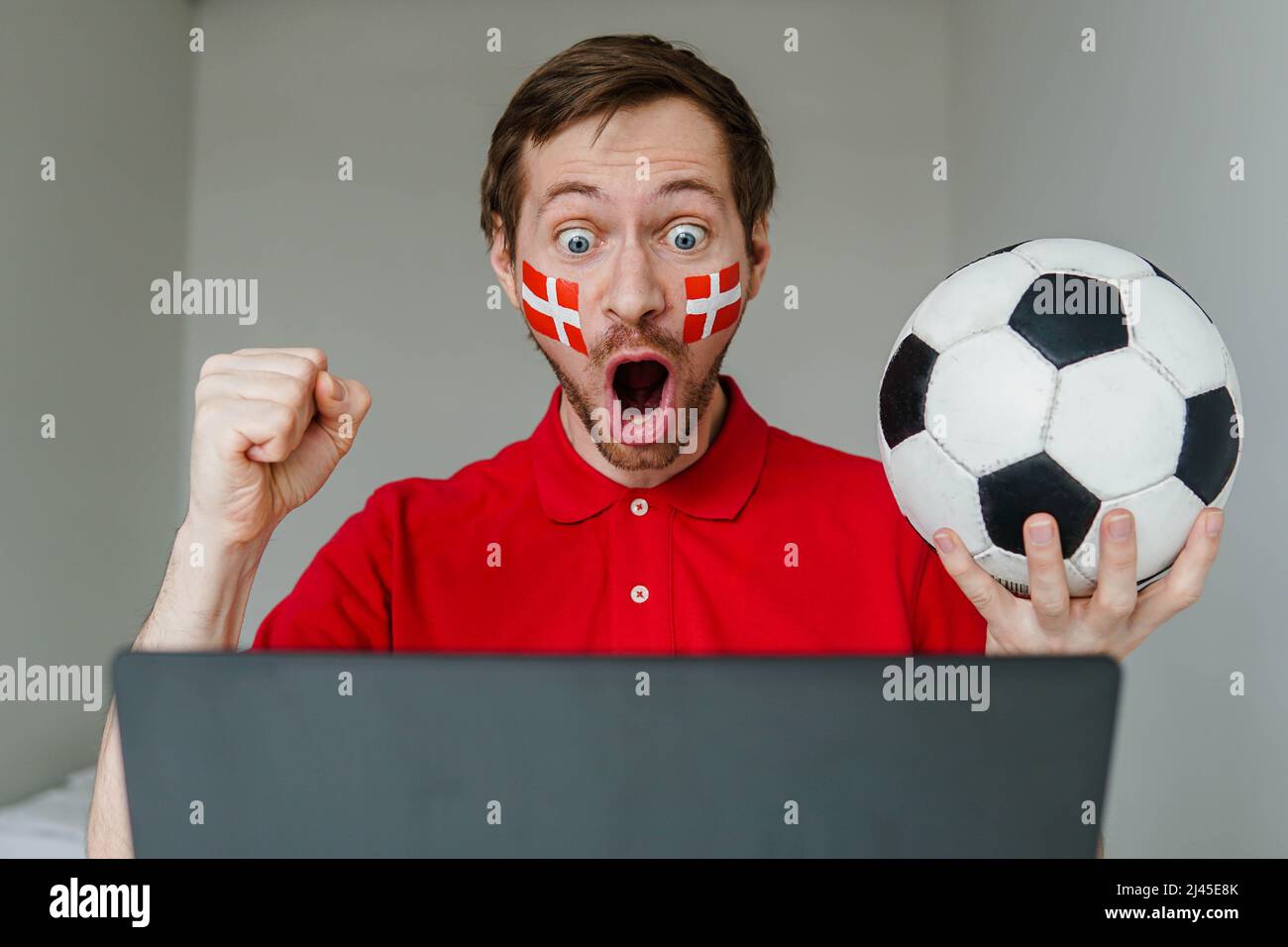 Junge aufgeregt Mann Fan unterstützen Dänemark Fußballnationalmannschaft halten in der Hand Fußball Fernsehen Live-Stream auf Laptop. Stockfoto