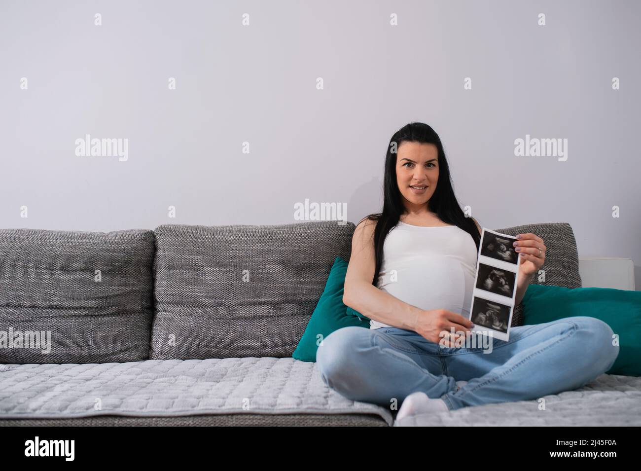 In fortgeschrittener Schwangerschaft sitzt die schwarze, langhaarige kaukasische Frau auf grünen Kissen gestützt.Schwangere mit riesigem Bauch zeigt das Ultraschallbild der Babys. Stockfoto